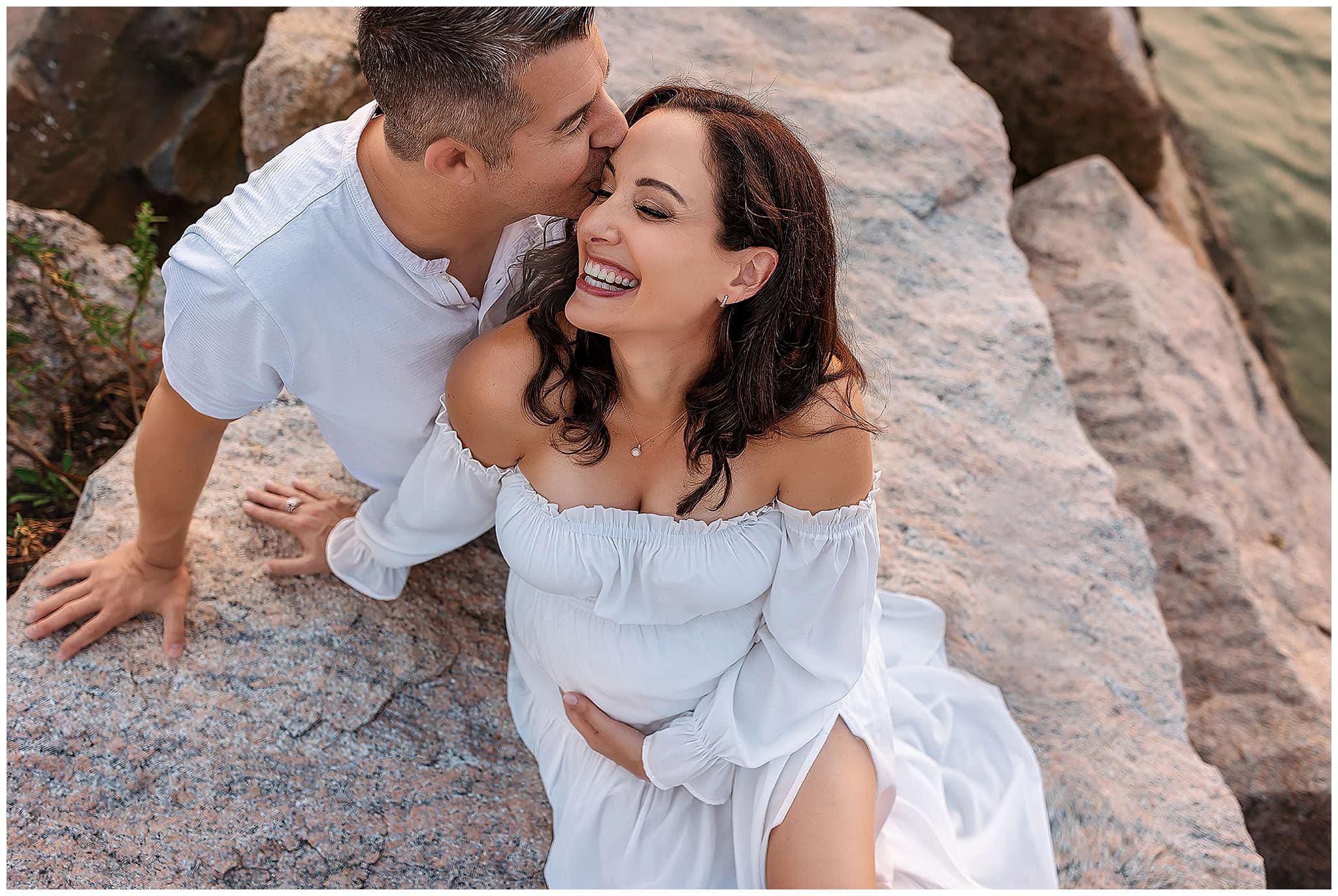 Nestled on a rocky Connecticut shoreline, the couple exudes pure joy. The man, clad in a white shirt, tenderly kisses the smiling woman on the forehead as she cradles her pregnant belly in a flowing white dress. Their expressions are serene and blissfully relaxed.