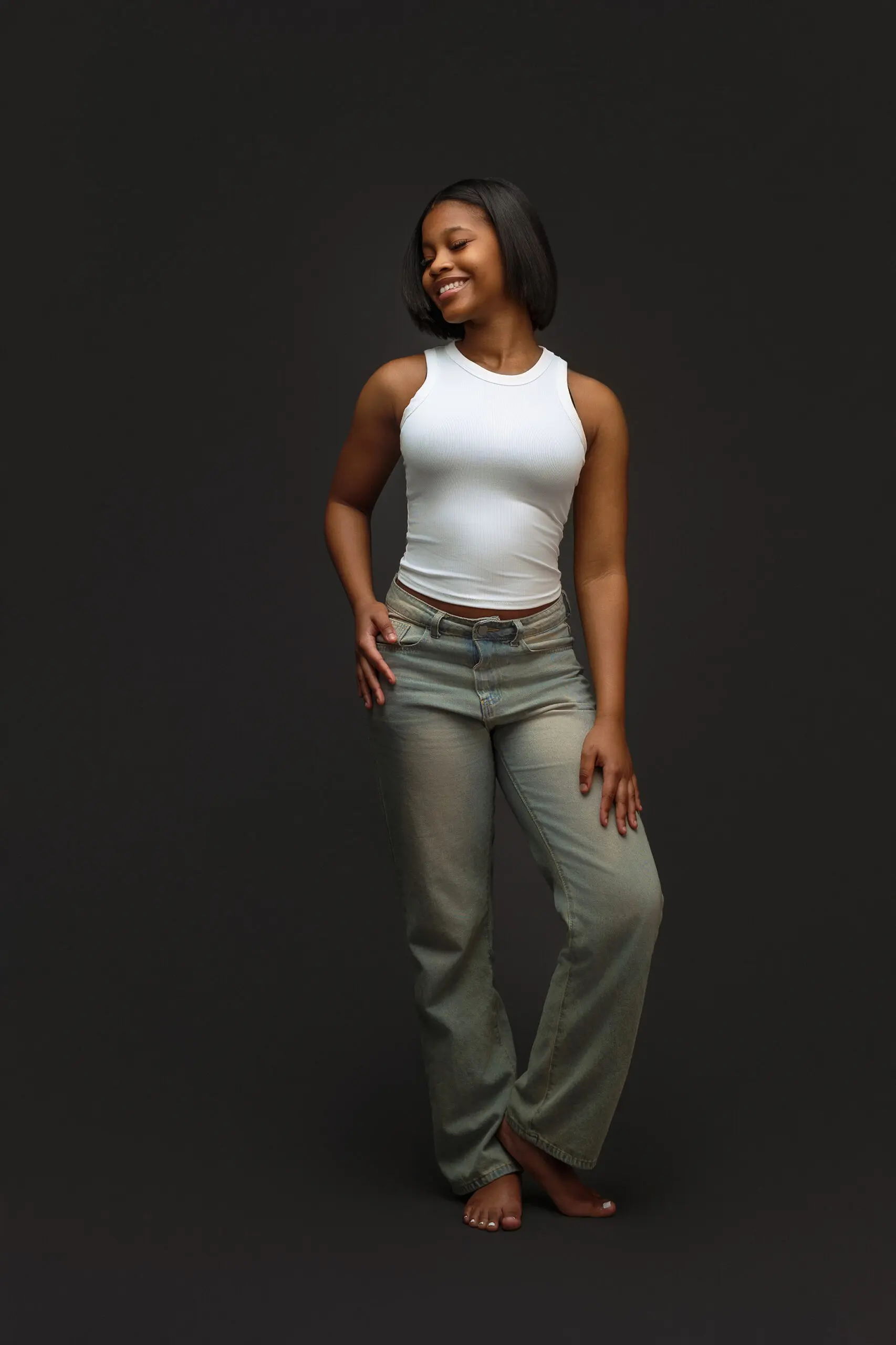 A woman stands barefoot against a dark background, smiling and looking to the side. She wears a sleeveless white top and light blue jeans with one foot crossed in front of the other.