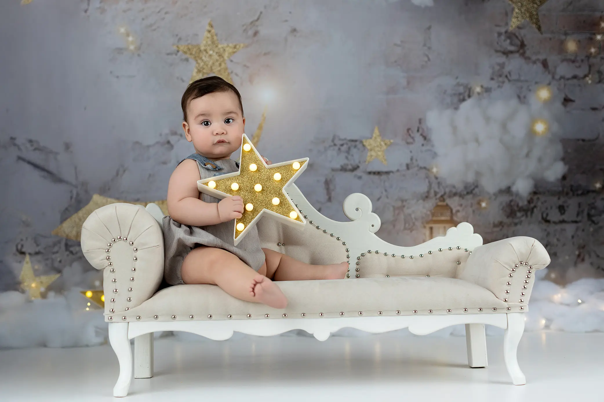 A baby sits on a small, elegant cream-colored couch, holding a lit-up gold star. The background features star decorations and fluffy clouds, creating a dreamy, whimsical atmosphere in a Connecticut studio.