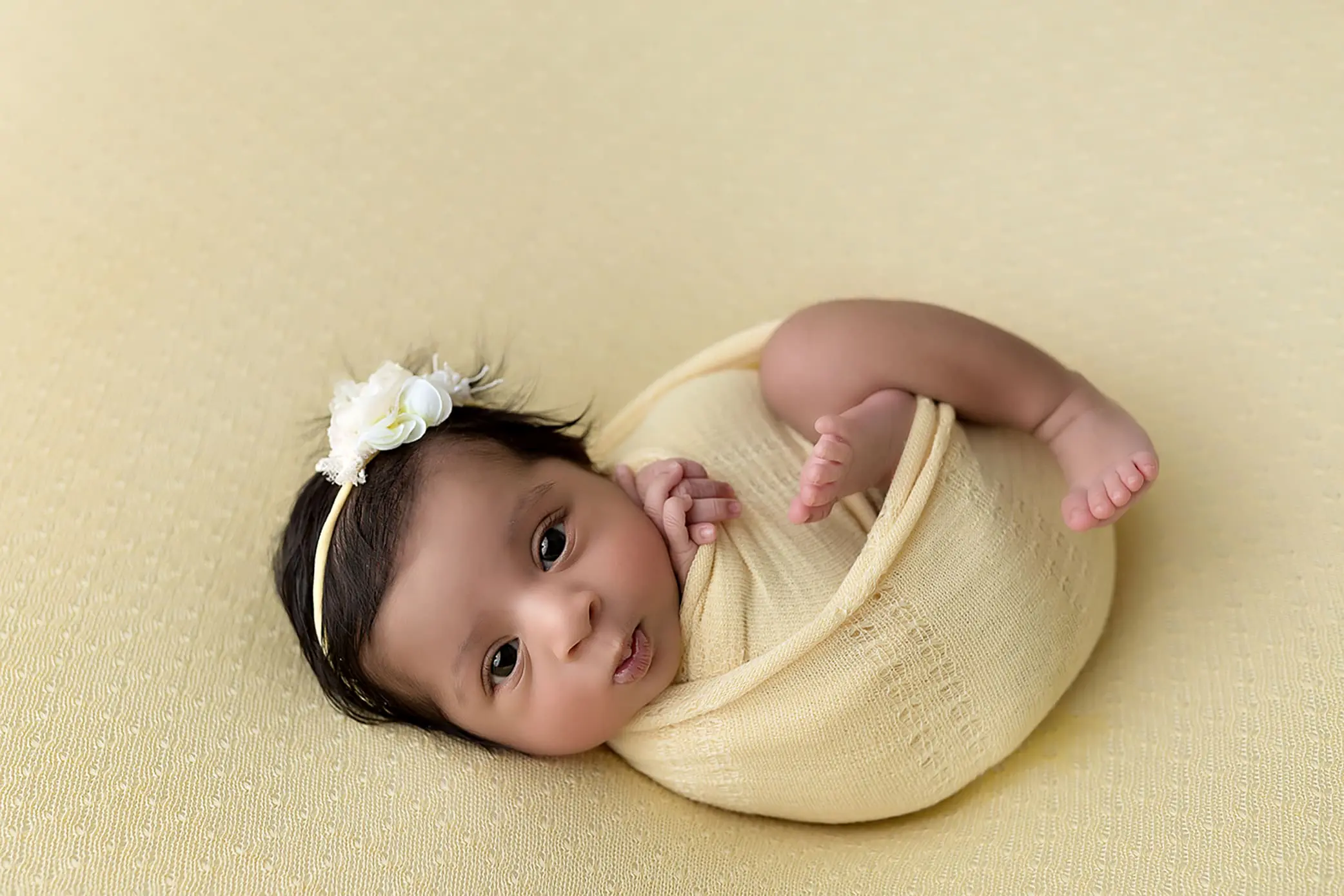 A baby is wrapped in a soft yellow blanket, lying on a matching yellow surface. With a white flower headband, the newborn gazes upward with calm eyes, tiny hands near their face and feet slightly visible—a perfect moment of newborn photography in Connecticut.