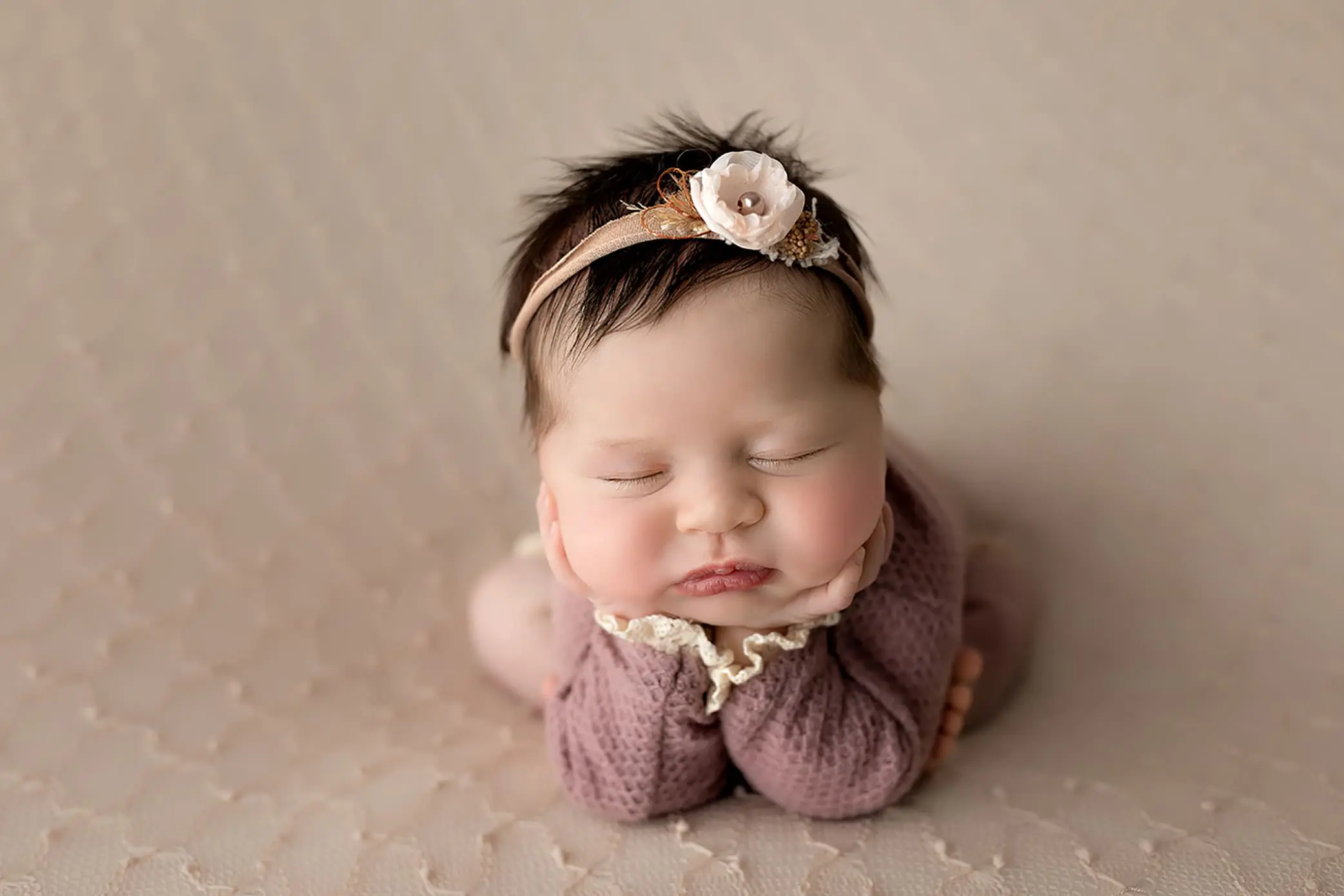 A sleeping baby with dark hair wears a pink headband with a flower, resting chin on hands, lying on a soft beige surface while dressed in a mauve knit outfit during her CT newborn photo session.