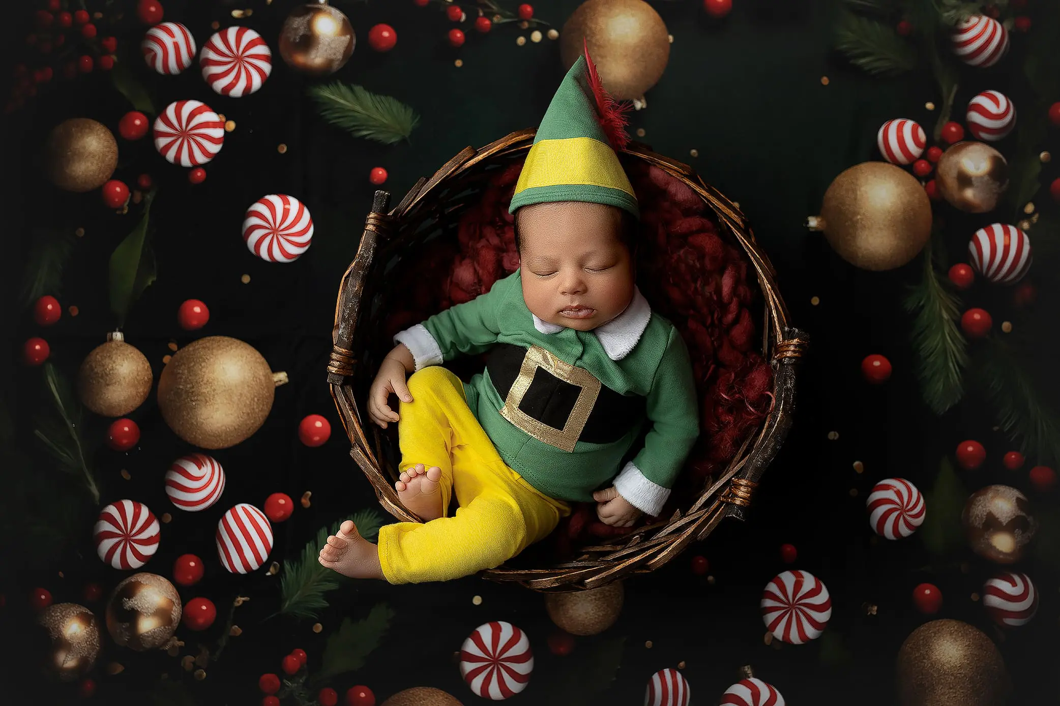A baby dressed as an elf sleeps peacefully in a woven basket, wearing a green top with a belt print, yellow pants, and a pointed hat. The basket is surrounded by festive decorations, including gold and striped ornaments with green foliage.