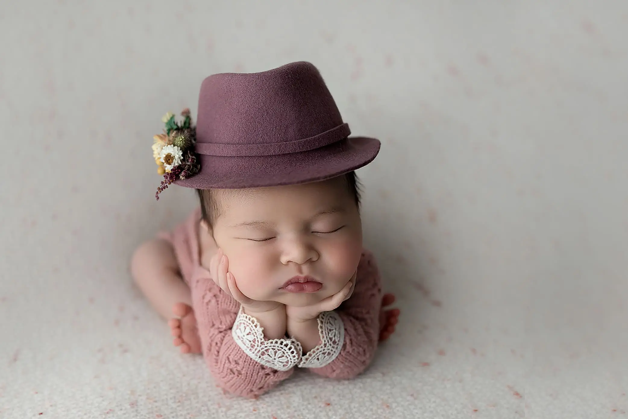 A sleeping baby rests on their stomach with their chin on their hands, captured beautifully by Connecticut’s trusted baby photographer. They wear a burgundy hat adorned with small flowers and a pink outfit with lace details. The background is softly textured in a light color.
