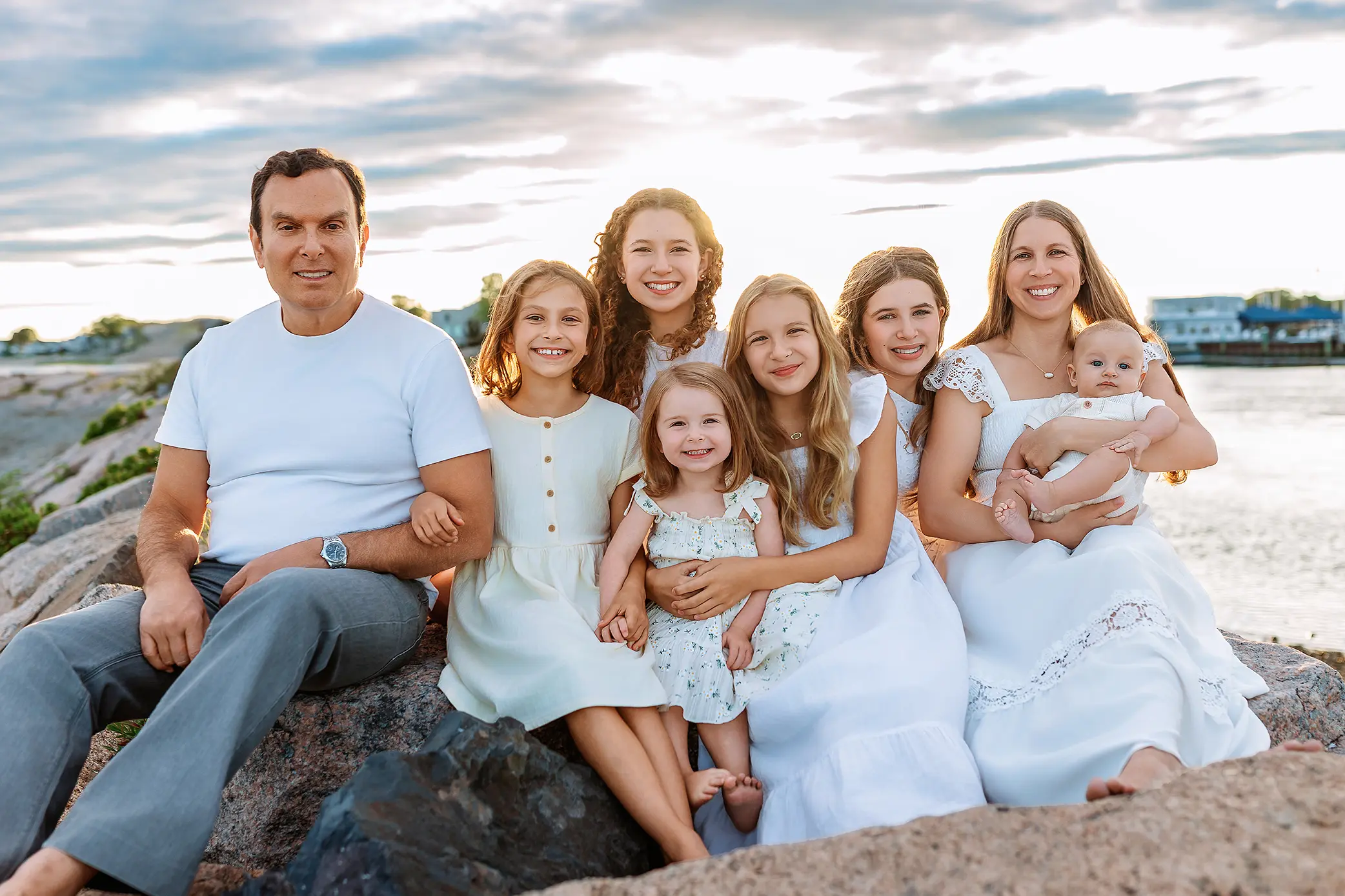 A family of seven, including one man, five girls, and one woman holding a baby, sit on rocks by the water at sunset, all smiling and dressed in light summer clothes during their CT family photo session.