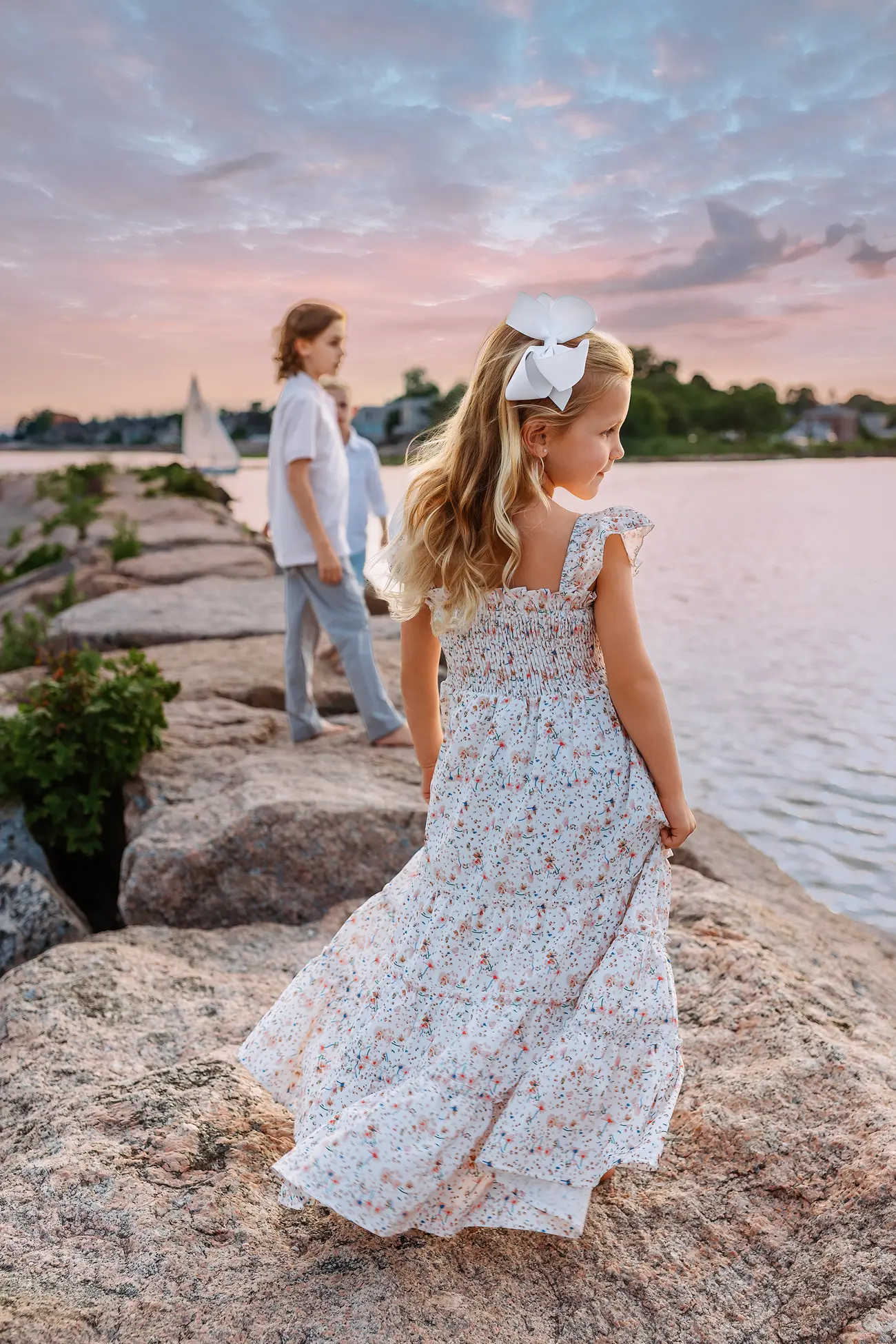 A young girl in a floral dress with a white bow stands on rocky shore, facing a boy and girl by the water at sunset. The sky is pink and blue, with trees and a sailboat in the background.