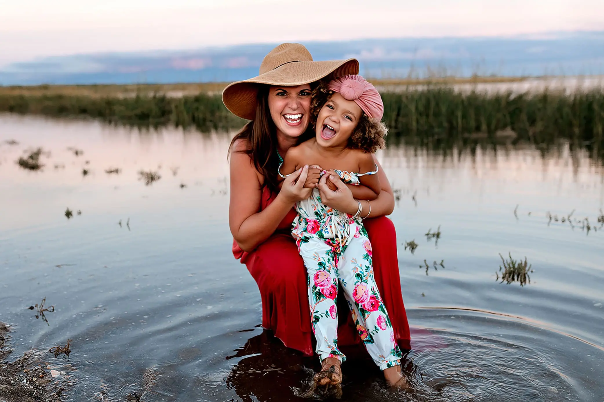A woman in a red dress and wide-brimmed hat kneels in shallow water with a smiling child wearing floral overalls and a pink headband. Their laughter echoes genuine, authentic family connection, surrounded by marshy grass and a serene body of water in the background.