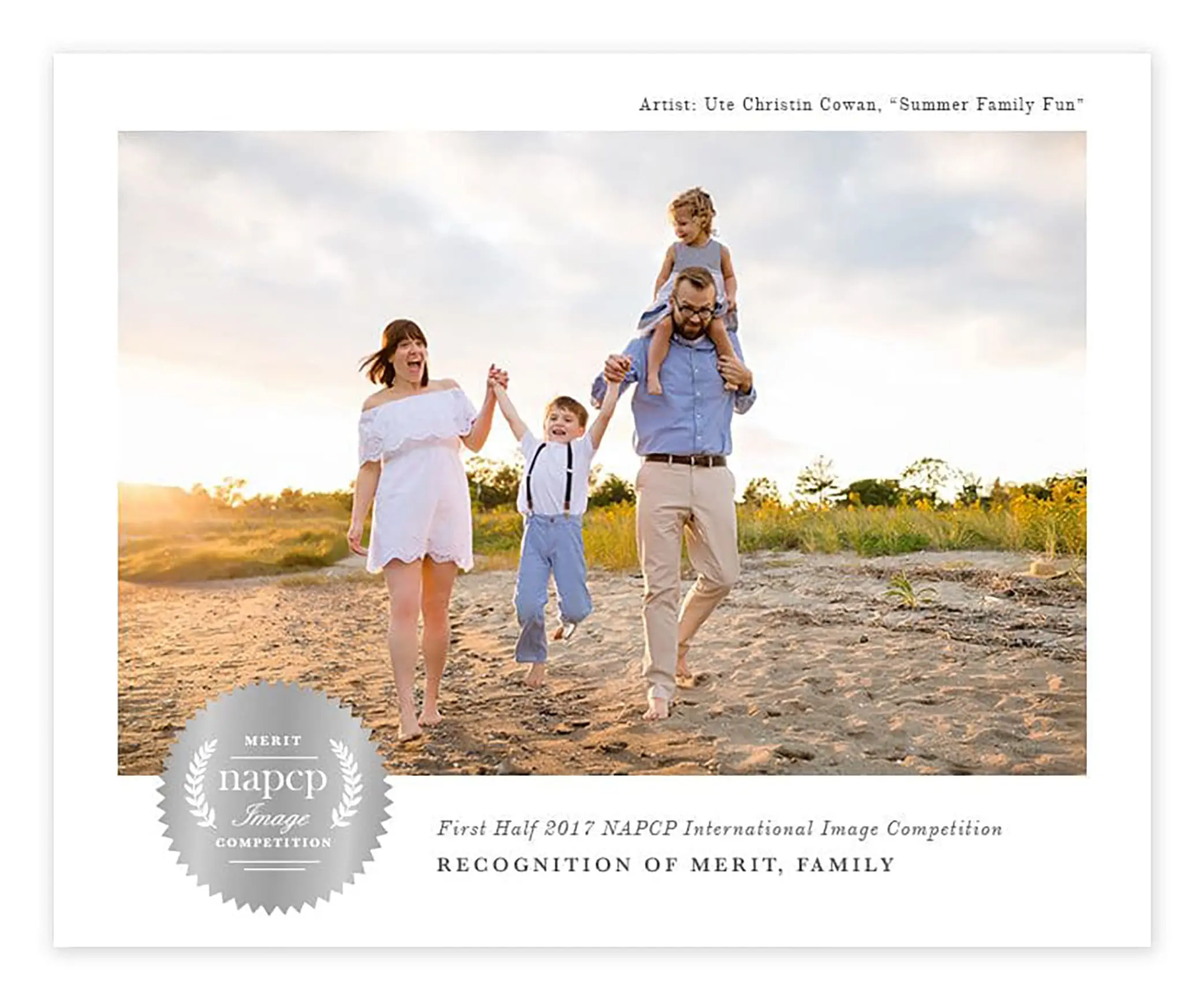 A family of four strolls along a sandy beach during their Connecticut family photography session. The father joyfully carries a child on his shoulders, while the mother, in a white dress, holds hands with another child. They smile under the partly cloudy sky—a scene worthy of the Merit NAPCP Image Competition badge.
