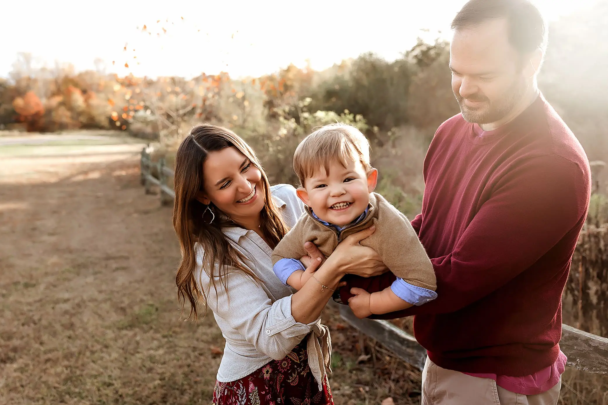 A woman and man stand outdoors in Connecticut, smiling and holding a young child between them. The child, dressed in a light brown top, is laughing. The background features a rustic fence and autumn foliage under a bright, sunny sky—a perfect family portrait.
