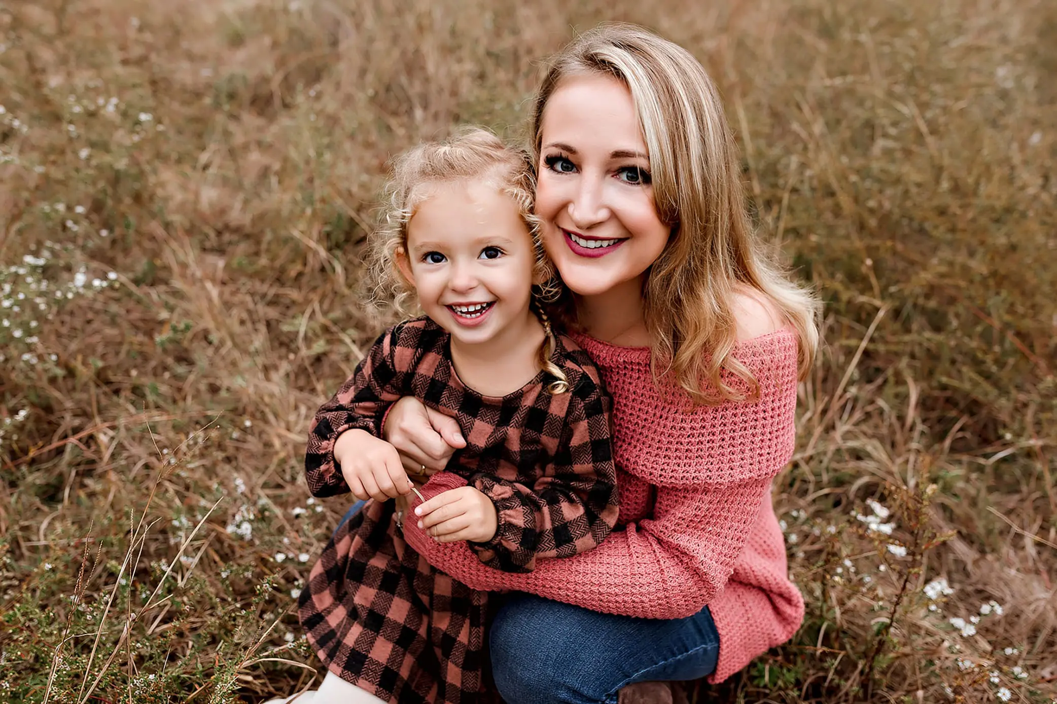 A woman in a pink sweater smiles while crouching beside a young girl in a checkered dress. They are enjoying one of the cherished family traditions in CT, taking an annual photo, sitting together in a grassy field dotted with small white flowers photographed by a CT family photographer.