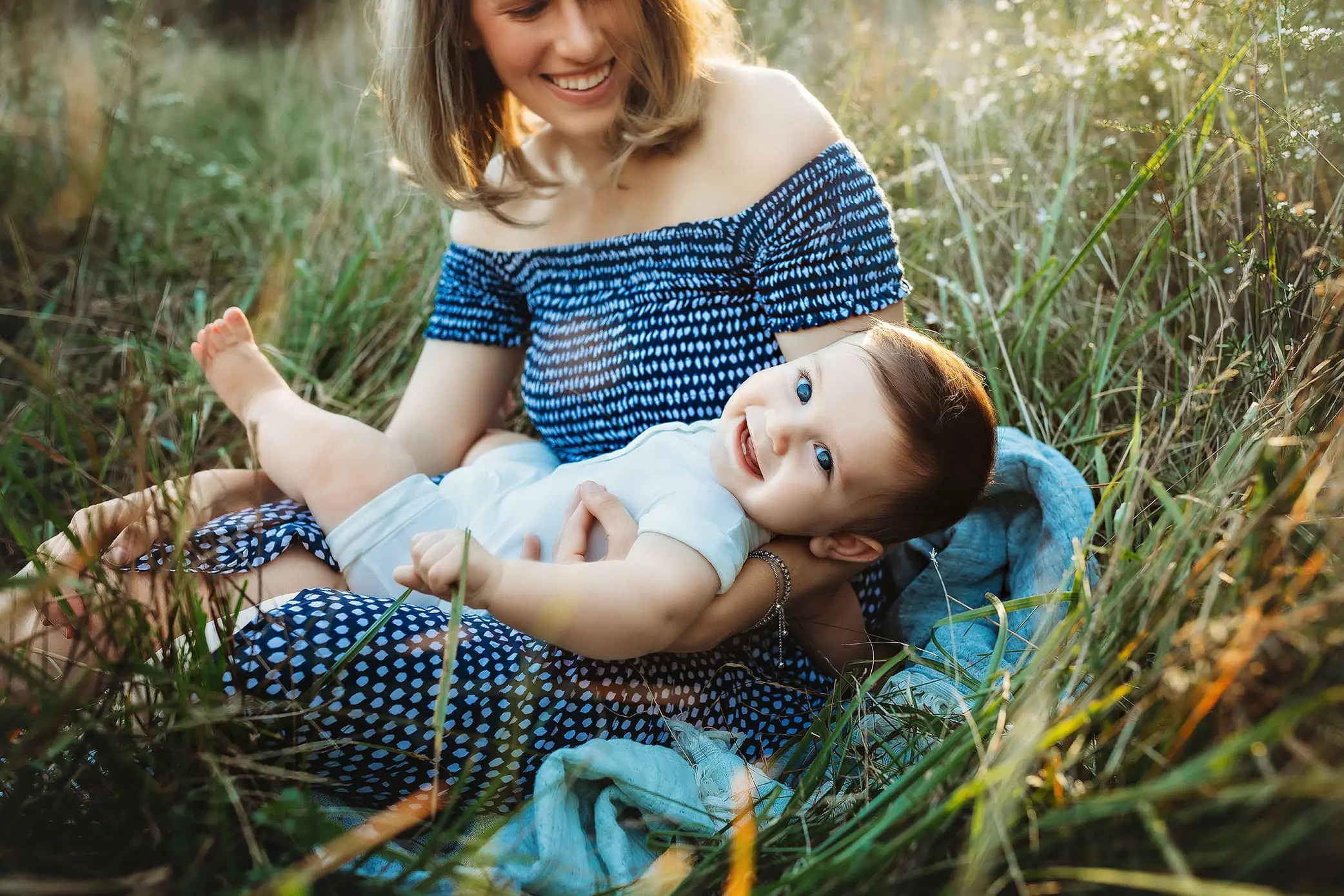 A woman sits in a grassy field wearing a blue patterned dress, smiling at a baby lying in her lap. The baby, in a light-colored outfit, looks up with a cheerful expression. Sunlight softly illuminates the scene.