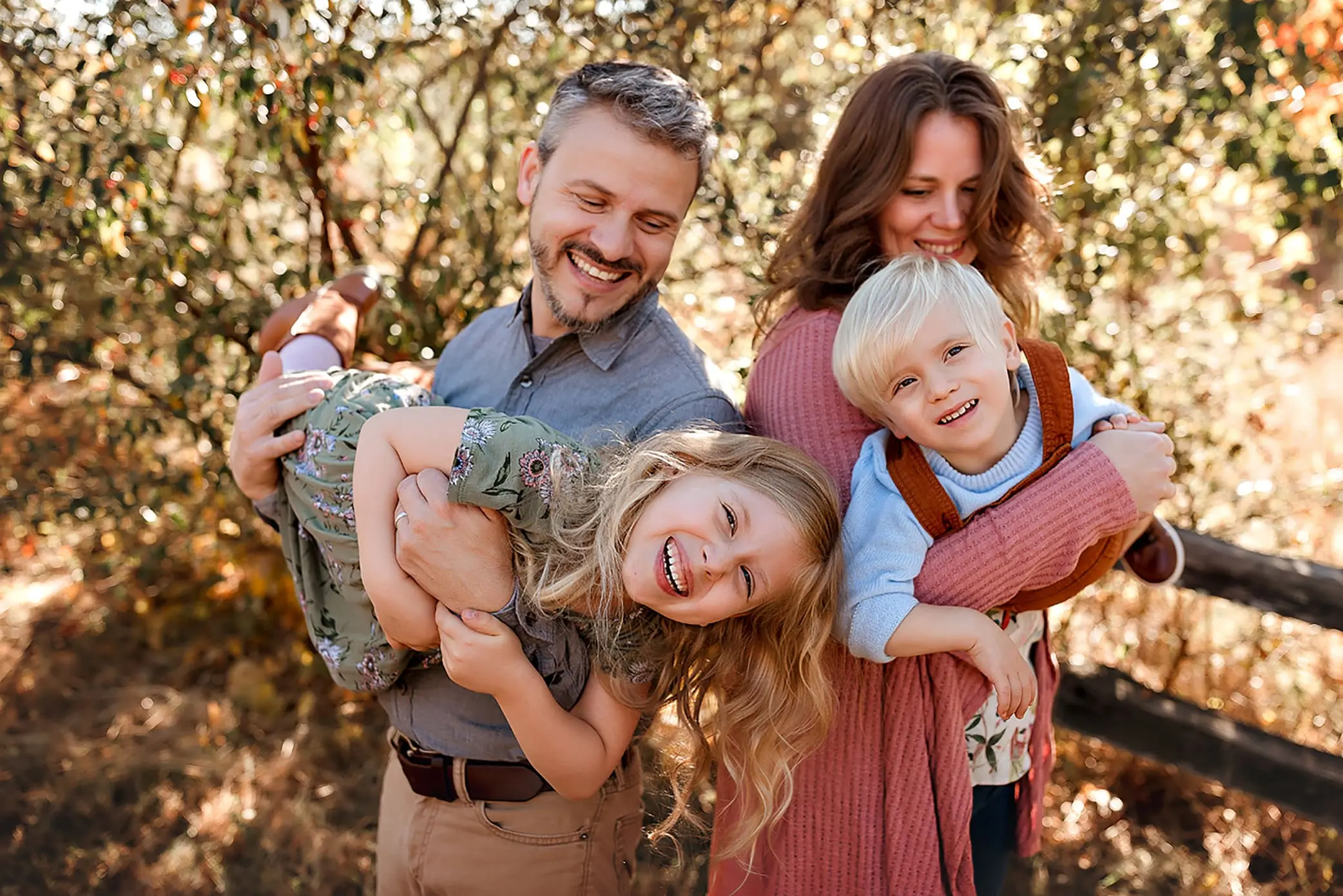 A smiling family of four outdoors. The father holds a laughing girl upside down, while the mother carries a smiling boy on her back. There are trees and sunlight in the background.