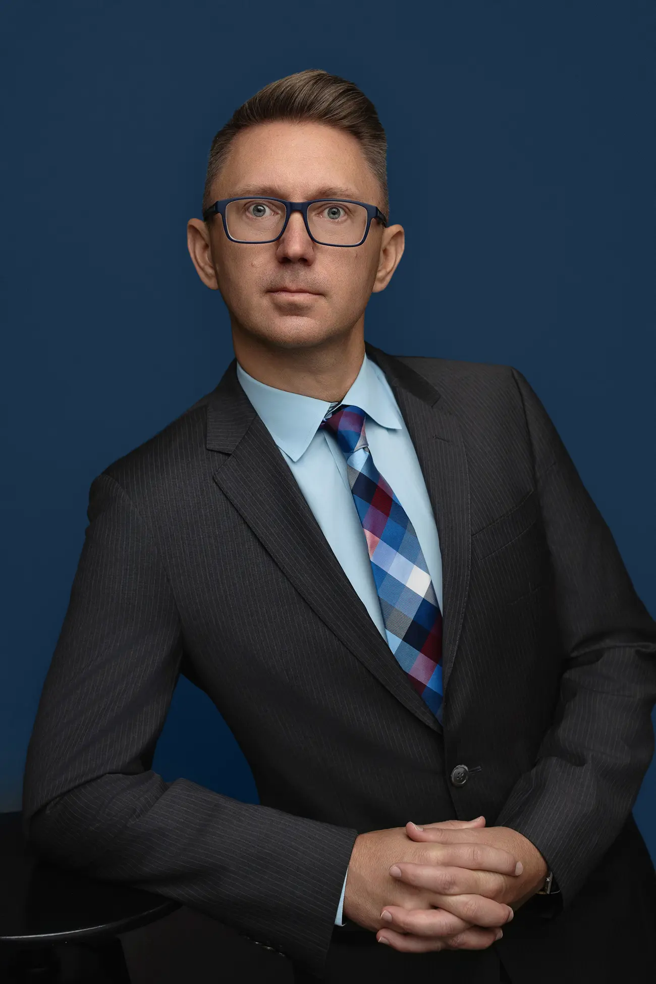 A man in a dark suit, light blue shirt, and patterned tie poses against a dark blue background, resting his hands on a black surface, looking slightly to the side with a serious expression.