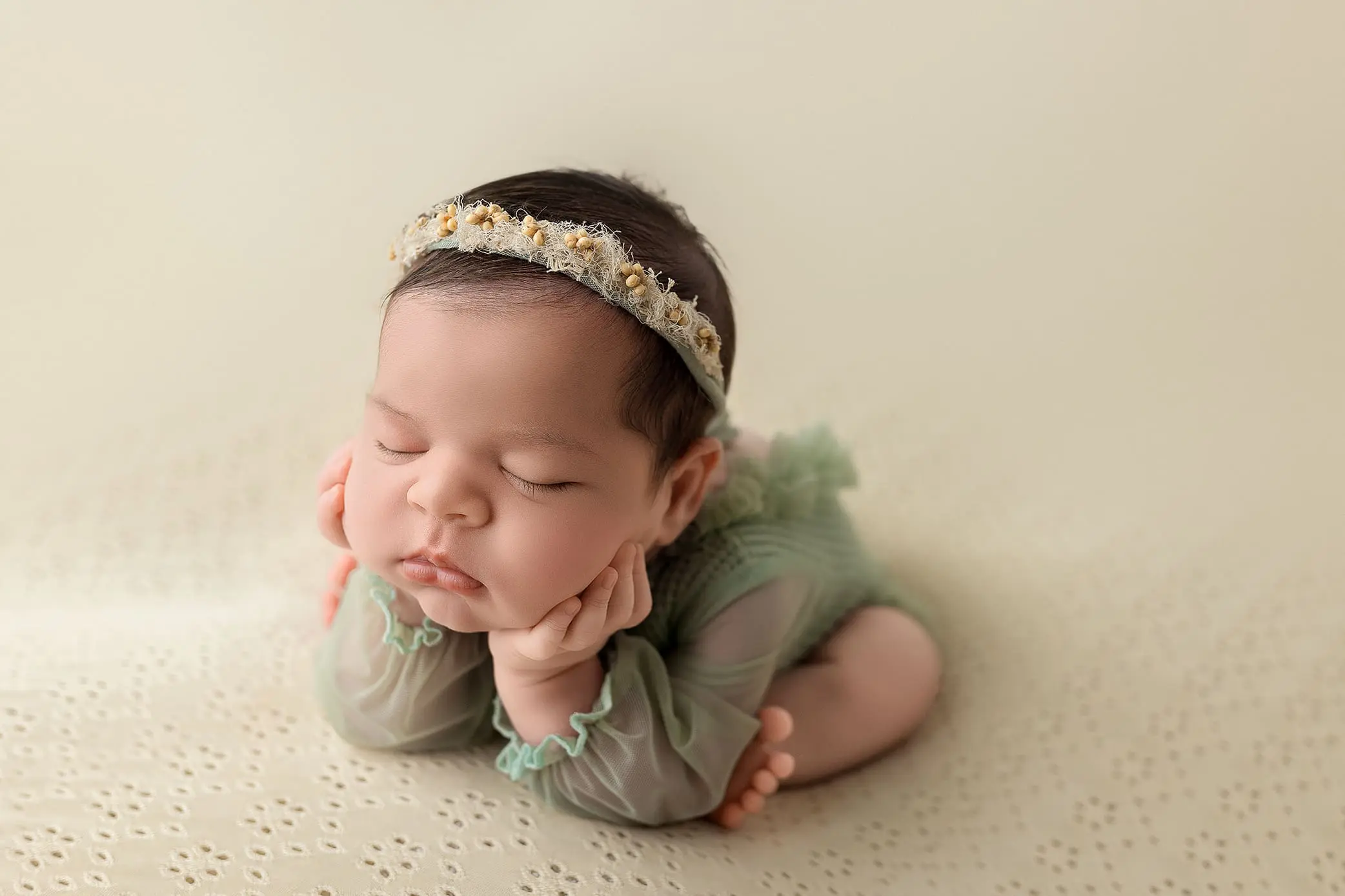 A sleeping baby lies on a soft, cream-colored surface. The baby wears a delicate green outfit and a floral headband. Their head rests on their hands, creating a peaceful and serene pose during her CT newborn session.