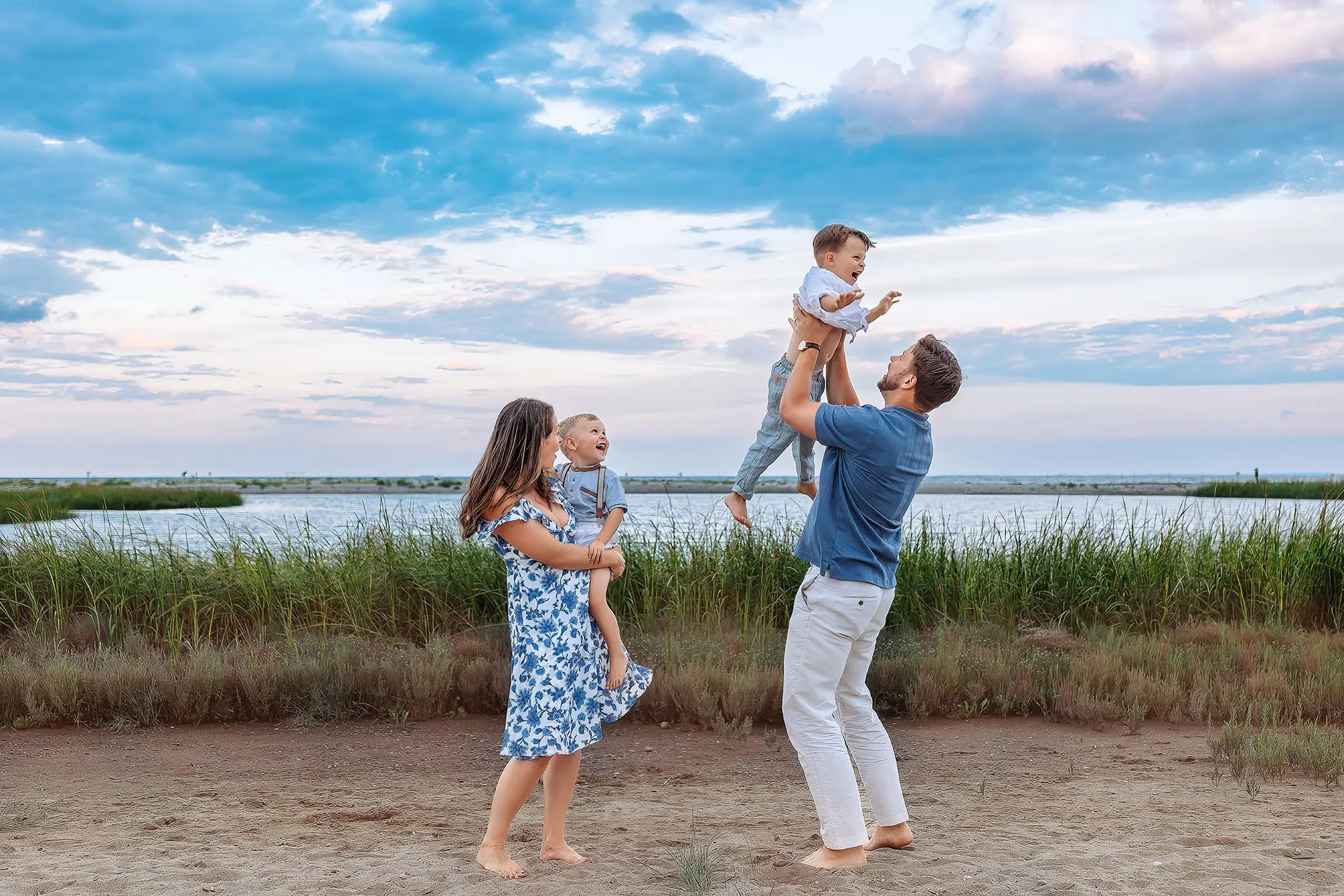 A family enjoys time at the beach. A man lifts a young child above his head, while a woman holds another child who is laughing. The sky is partly cloudy, and the water and grass create a serene background for a family photo shoot in Connecticut.