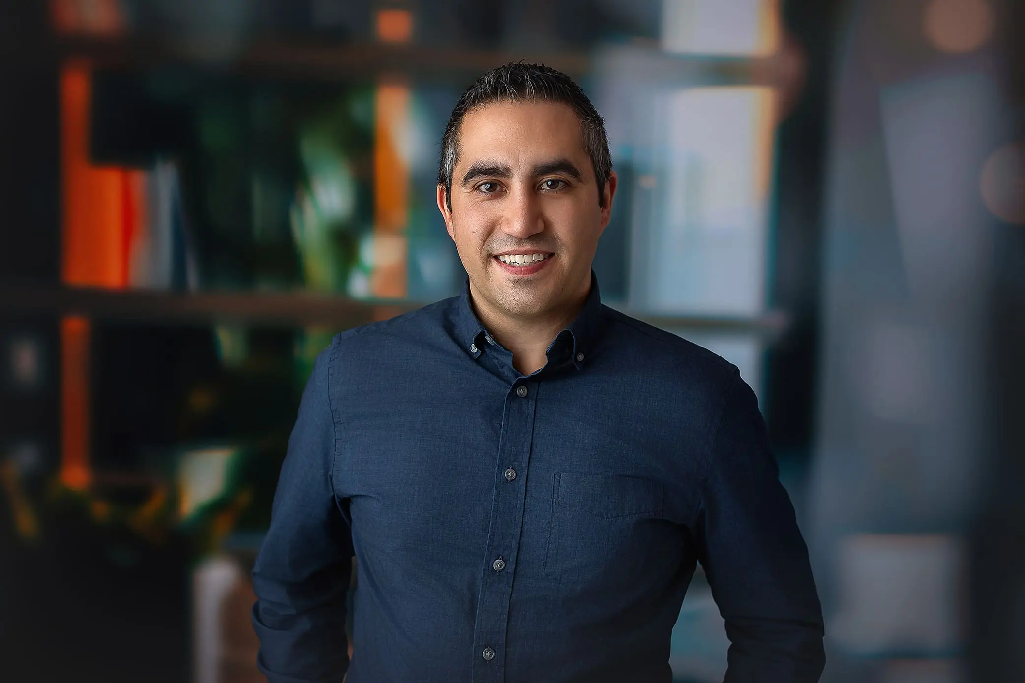 A man in a dark blue button-up shirt is smiling at the camera. He stands in front of a blurred background featuring shelves with books and plants posing for a CT Headshot. Captured by Headshot Photographer in Connecticut.