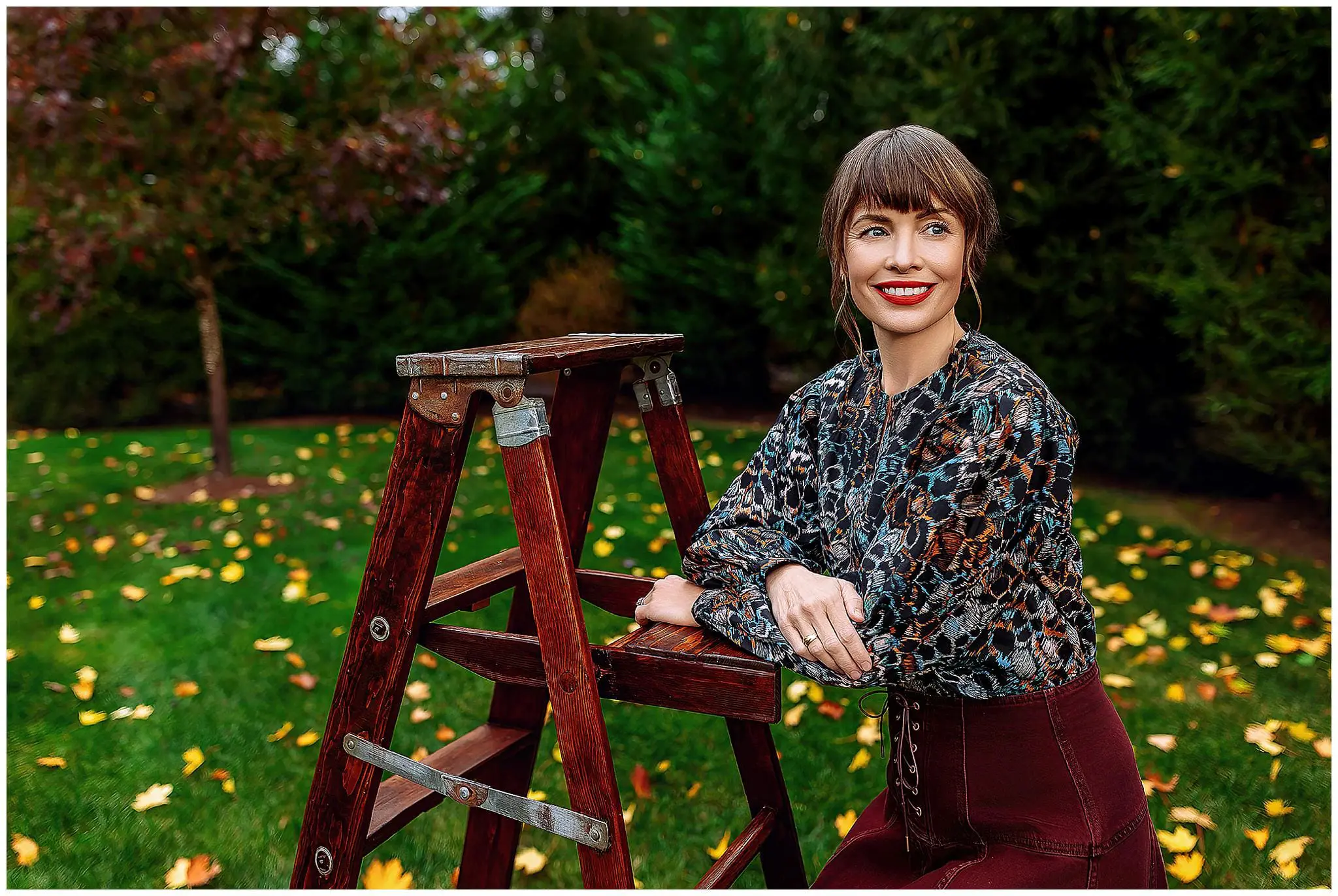 A woman with short brown hair, wearing a patterned blouse and maroon skirt, leans on a wooden ladder outdoors. The ground is scattered with yellow leaves, and trees with green and red foliage are in the background. She is smiling.