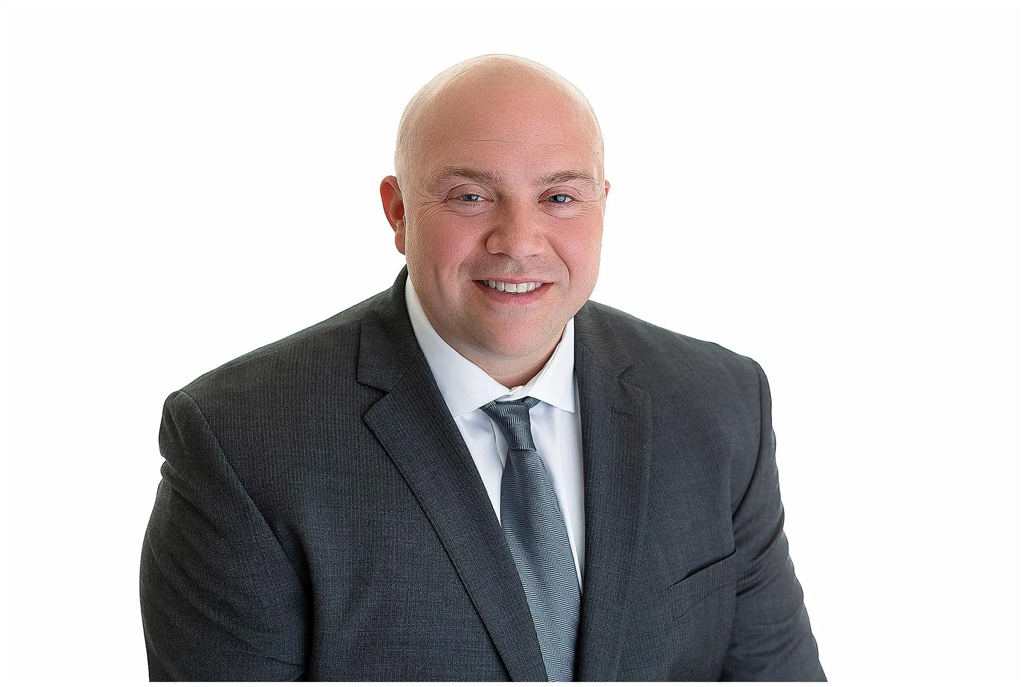 A bald man in a dark suit and blue tie is smiling at the camera against a plain white background.