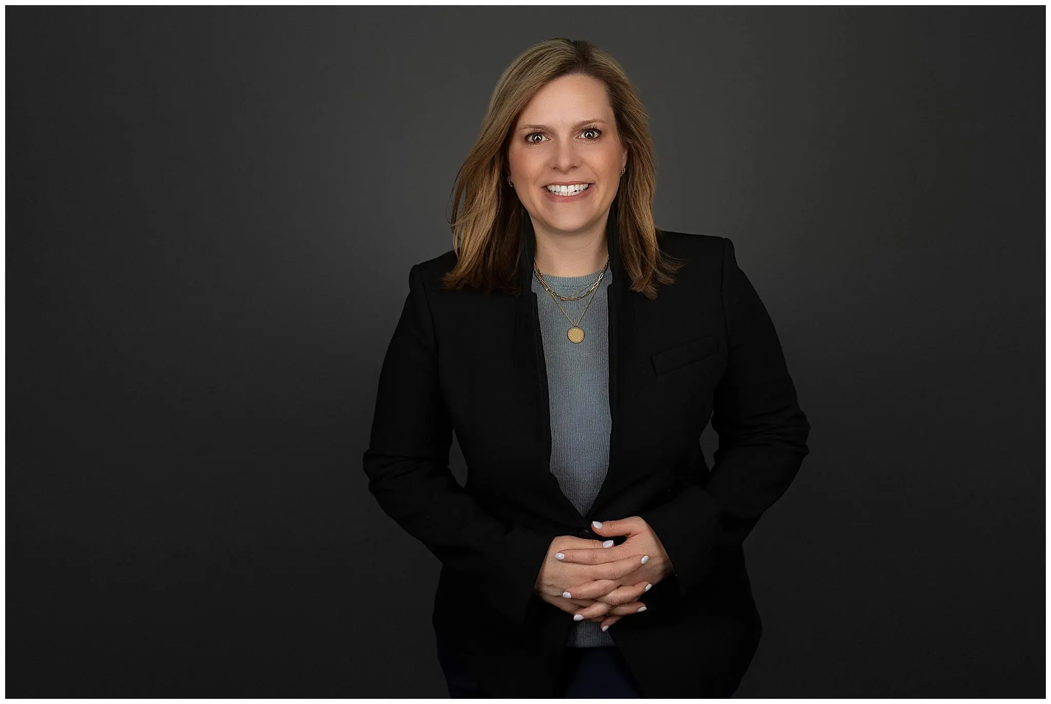 A woman with shoulder-length brown hair, wearing a black blazer over a gray top, smiles at the camera in this professional headshot. She is standing against a dark gray background with her hands clasped in front of her.