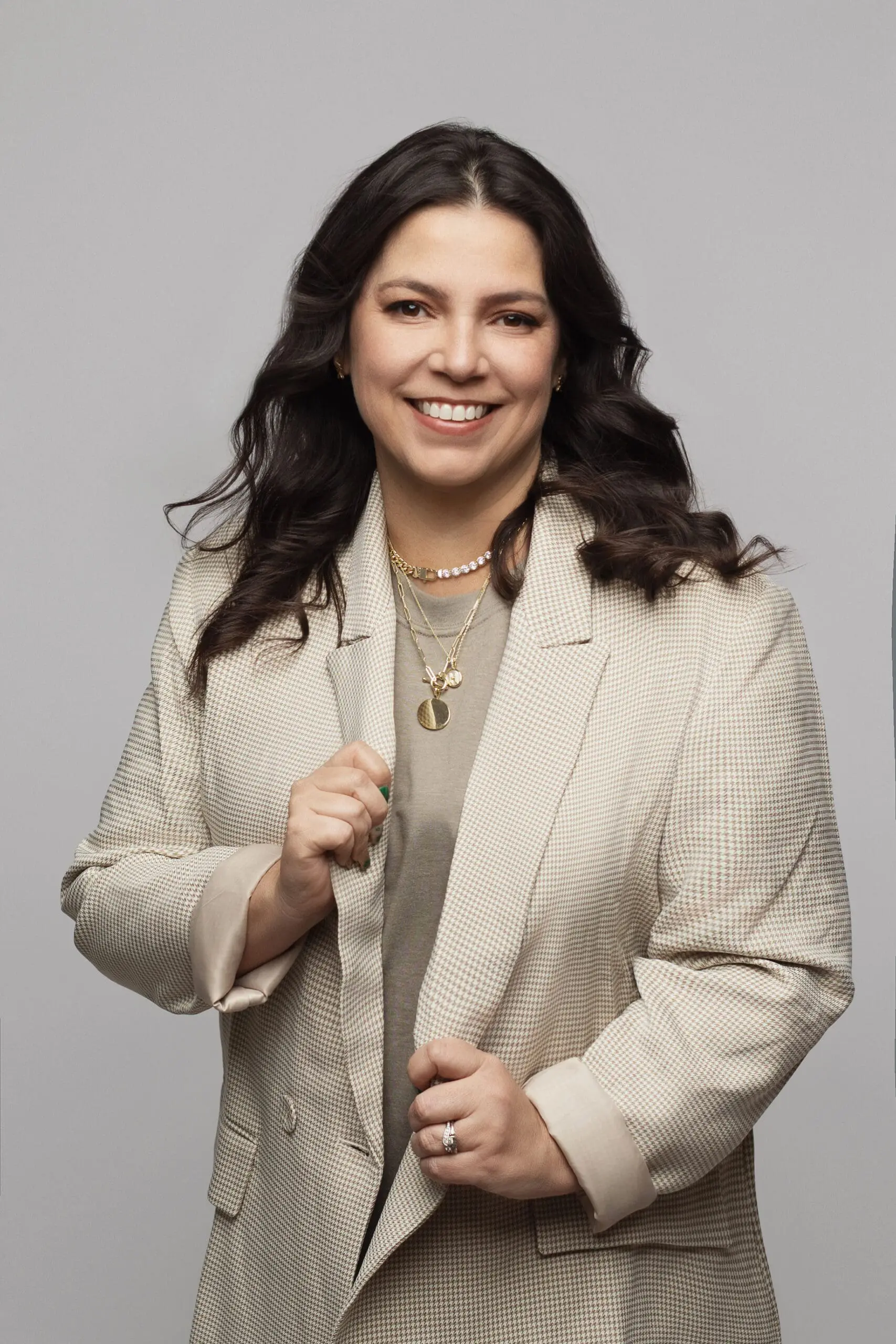 Smiling person with long dark hair wearing a beige blazer, taupe shirt, and layered necklaces against a plain gray background during her Connecticut Branding session.