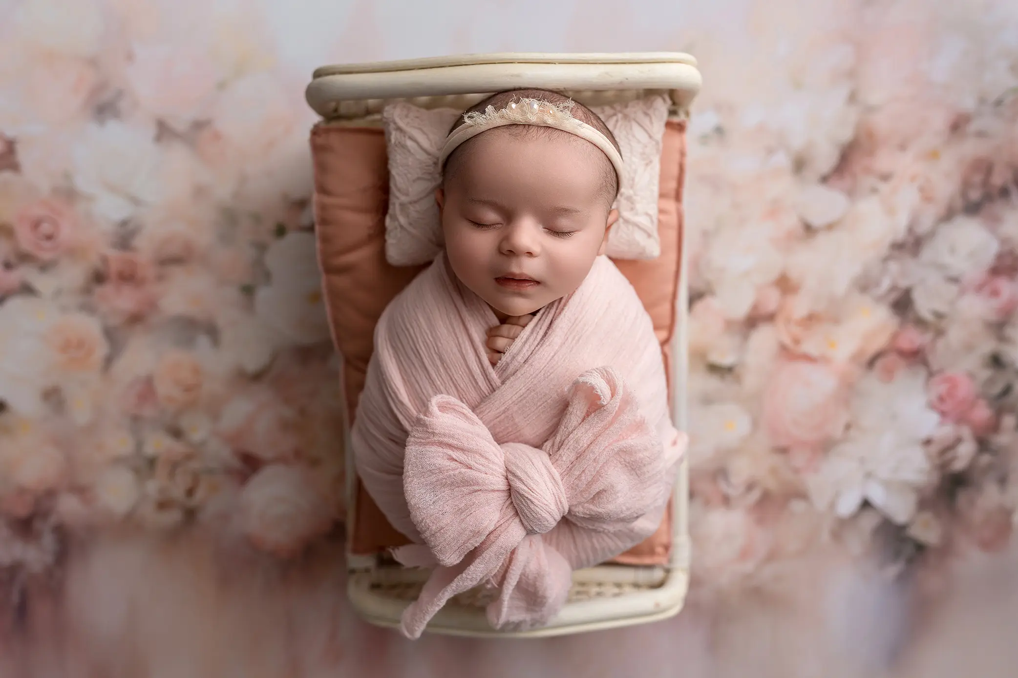 A newborn baby wrapped in a soft pink blanket with a large bow, lies asleep on a small bed with peach bedding and a floral background. The baby wears a delicate headband and smiles gently.