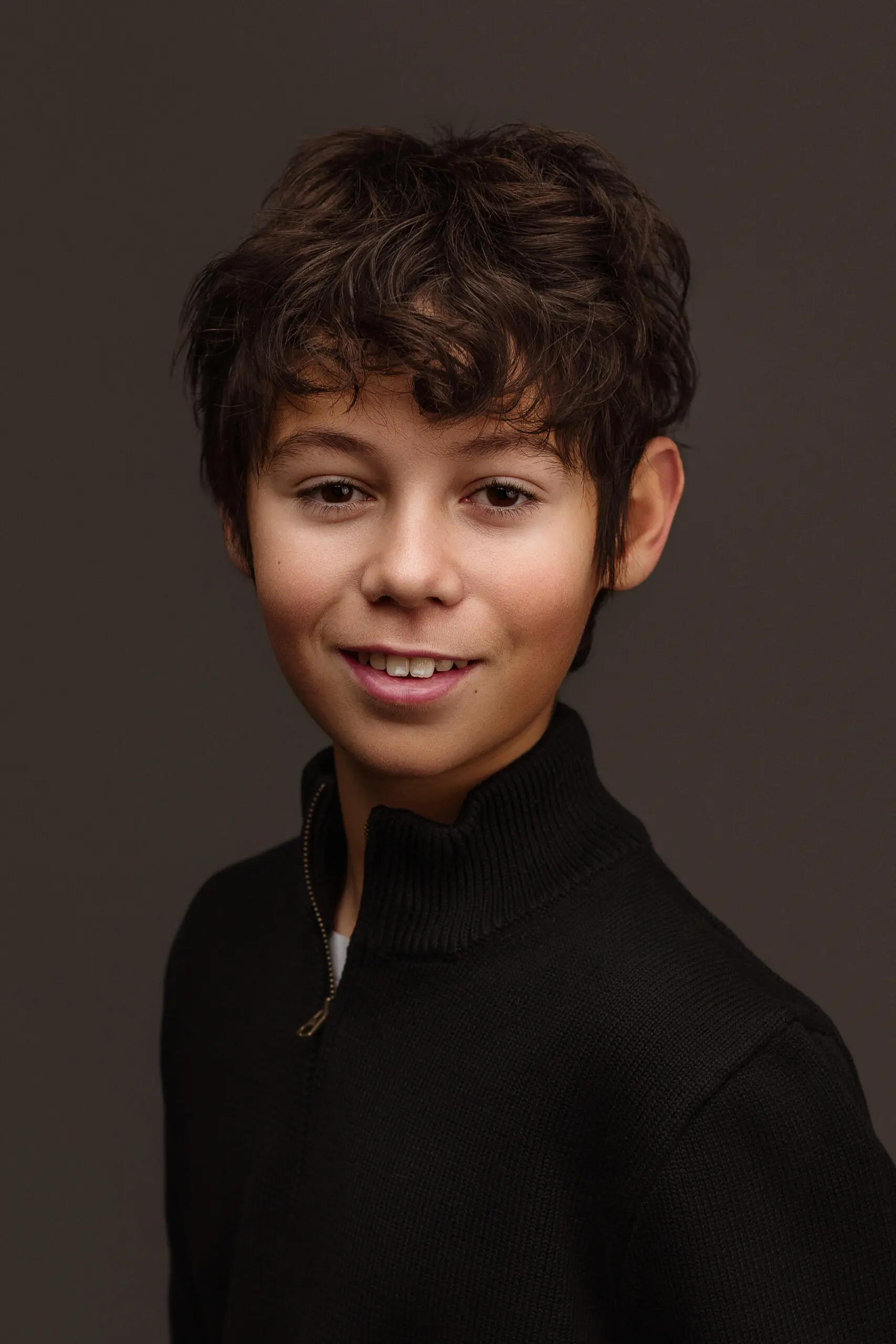 A young boy with wavy brown hair wearing a black zip-up sweater smiles gently in this headshot branding photo, posed against a plain dark background.