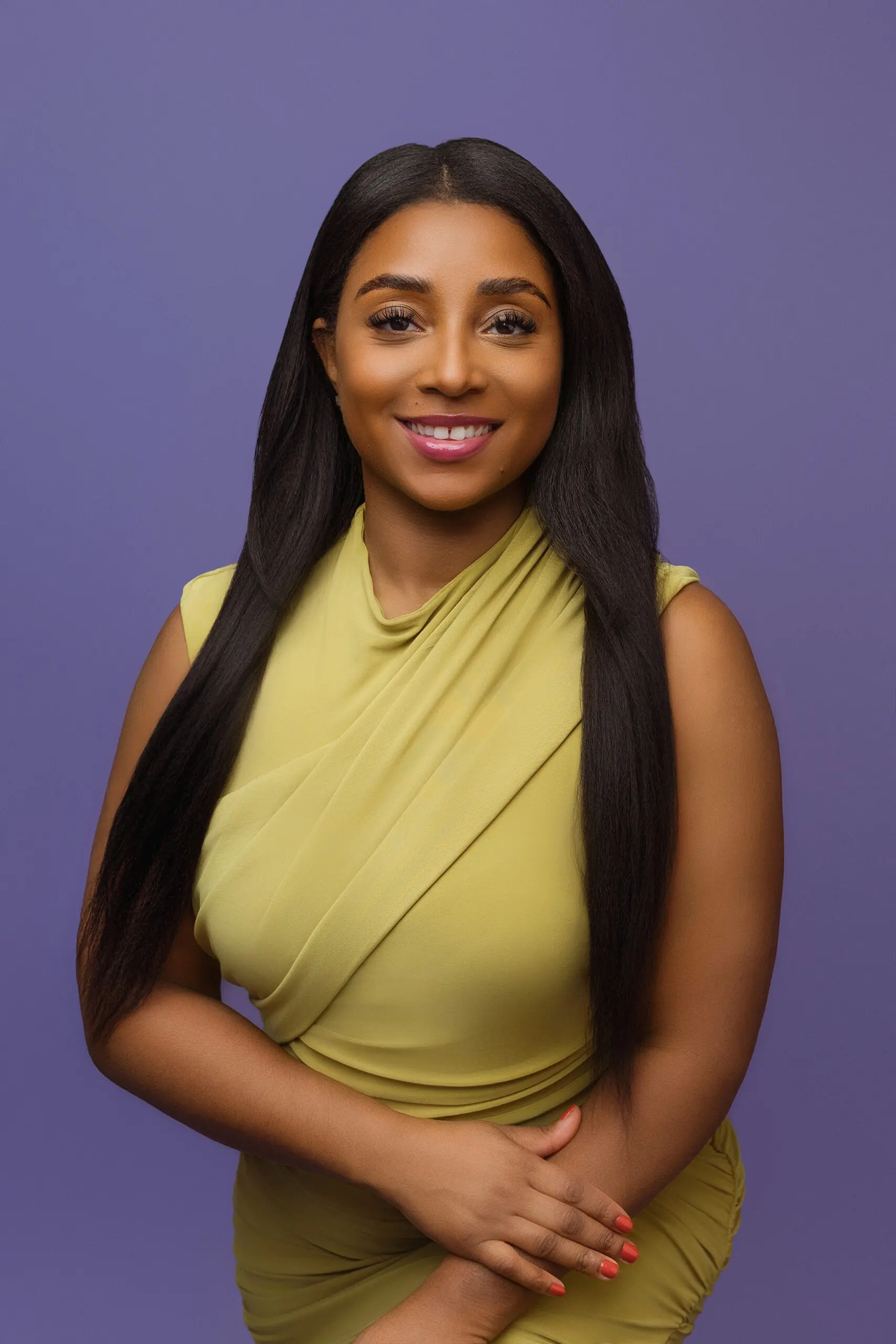 A woman with long straight dark hair wearing a sleeveless yellow dress smiles while posing with her arms crossed against a solid purple background.