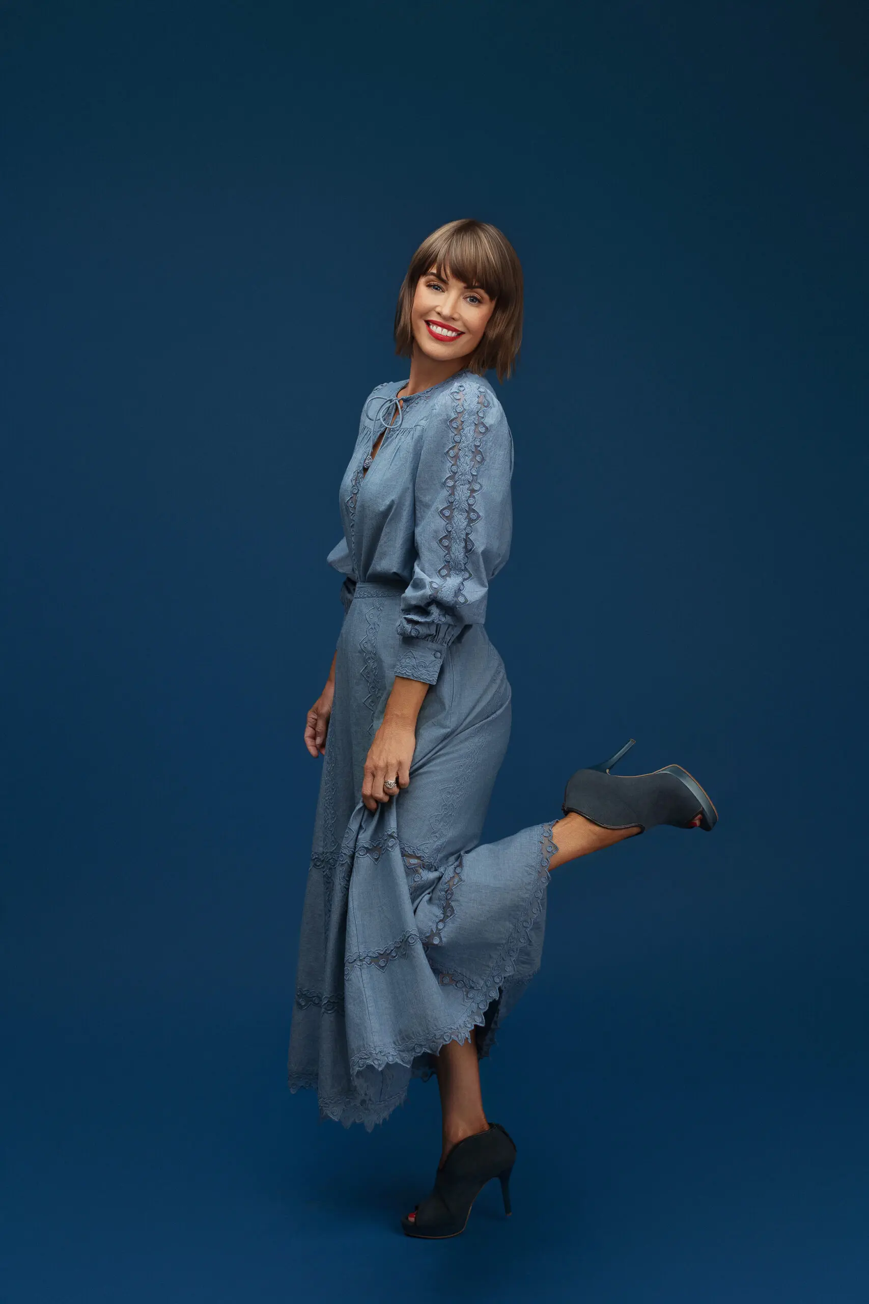 A woman in a light blue, long-sleeved dress stands against a dark blue background, smiling with one leg bent and lifted behind her. She wears dark high-heeled shoes and has short brown hair with bangs.