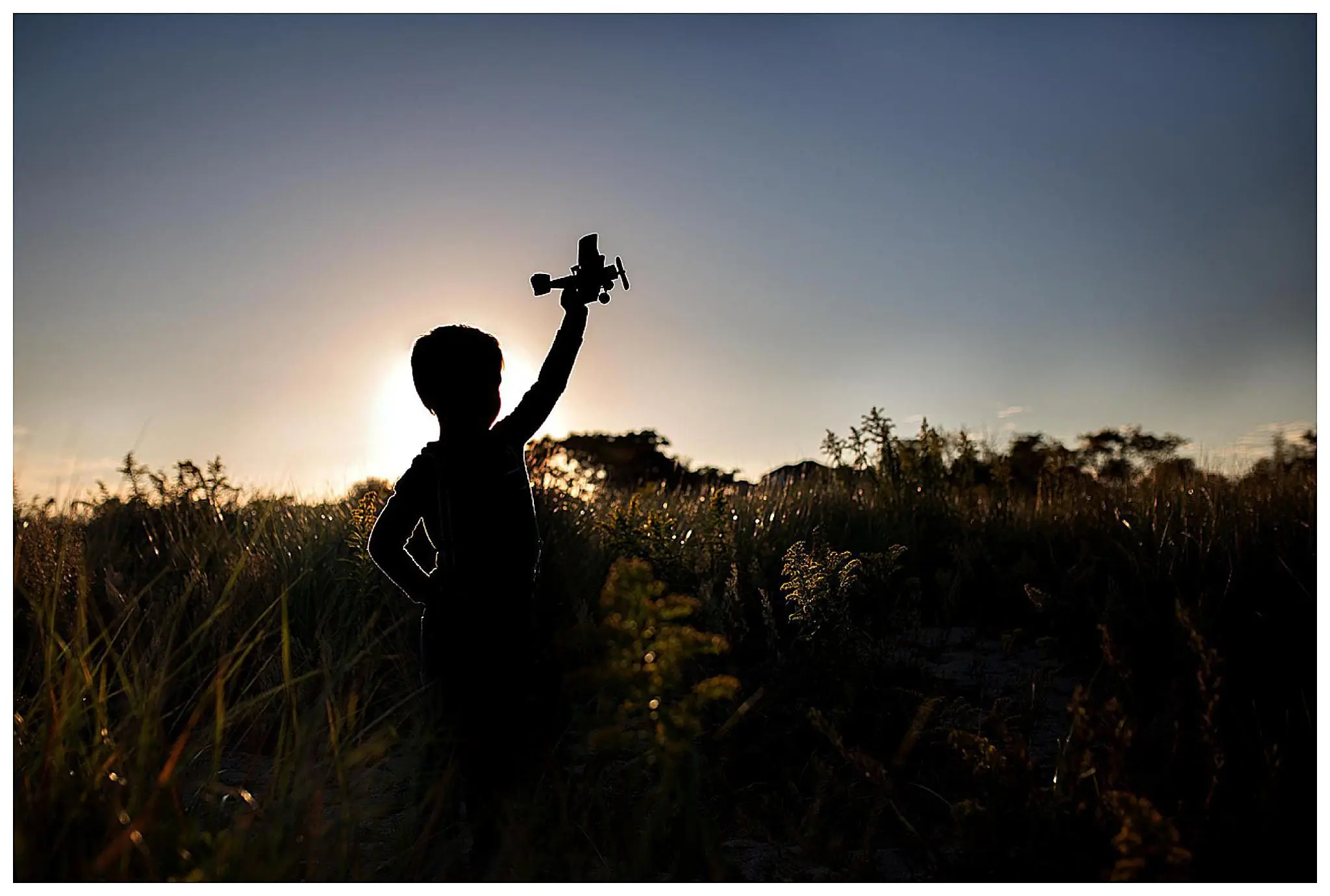 Silhouette of a child holding a toy airplane up against the setting sun, standing in a field with tall grass. The sky is clear, creating a peaceful and dreamy atmosphere.