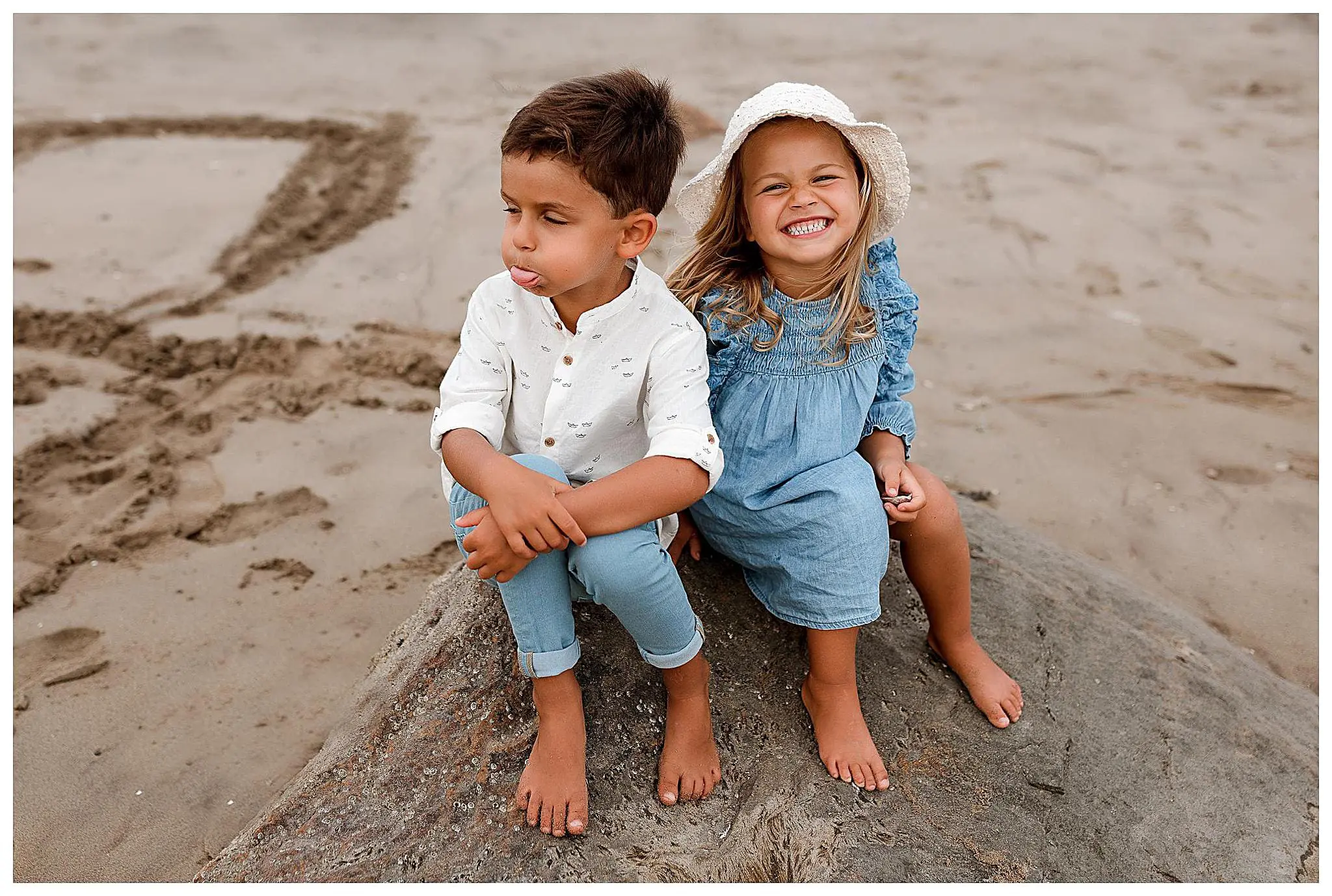 Two children sit on a large rock at the beach in Milford Connecticut. The boy, pouting and wearing a white shirt and blue pants, sits with his arms crossed. The girl wears a blue dress and white hat, smiling widely. Sand and beach are visible in the background.