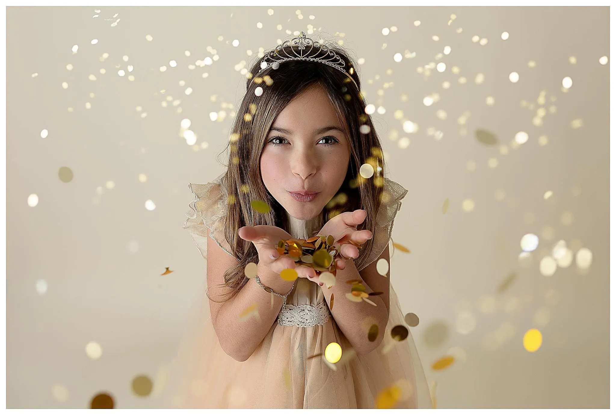 Young girl wearing a tiara and a light-colored dress blows a handful of gold confetti towards the camera. The background is a soft, neutral color with sparkling bokeh effects, creating a magical, celebratory atmosphere.