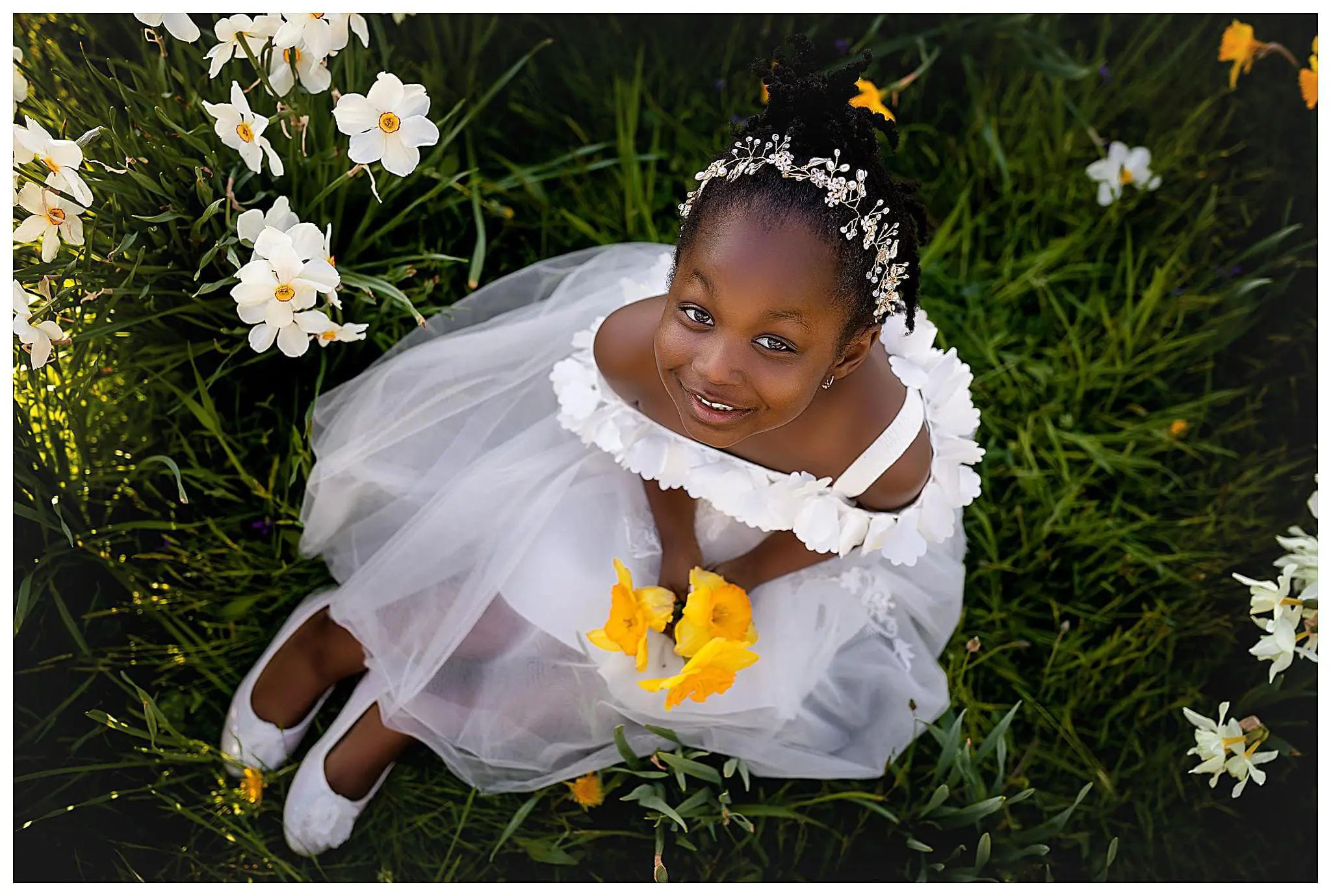 A young girl in a white dress sits on green grass with white flowers around her. She holds yellow flowers, smiling and looking up. Her hair is styled with a floral headband, and she wears white shoes.