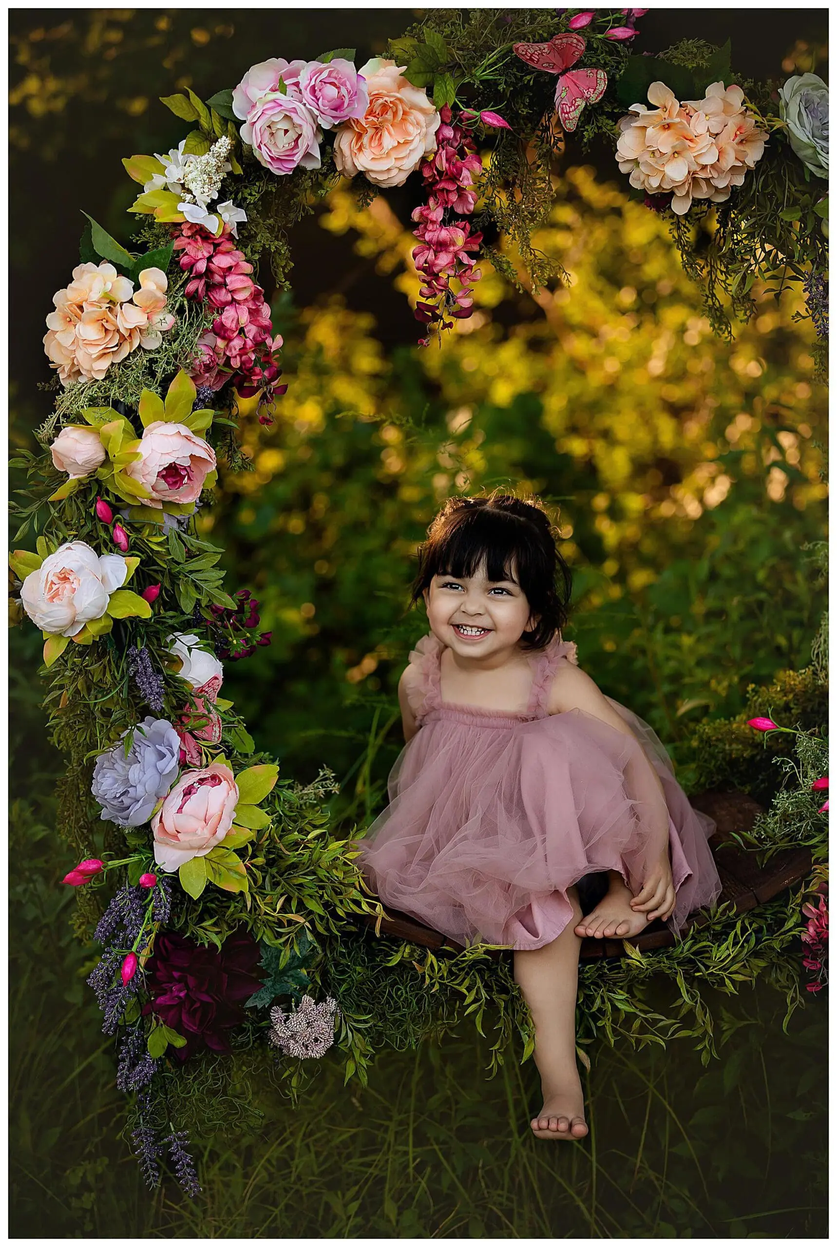 A young child in a pink dress smiles while sitting on a swing adorned with colorful flowers and greenery. The background is a lush green, creating a whimsical and enchanting scene in Connecticut.