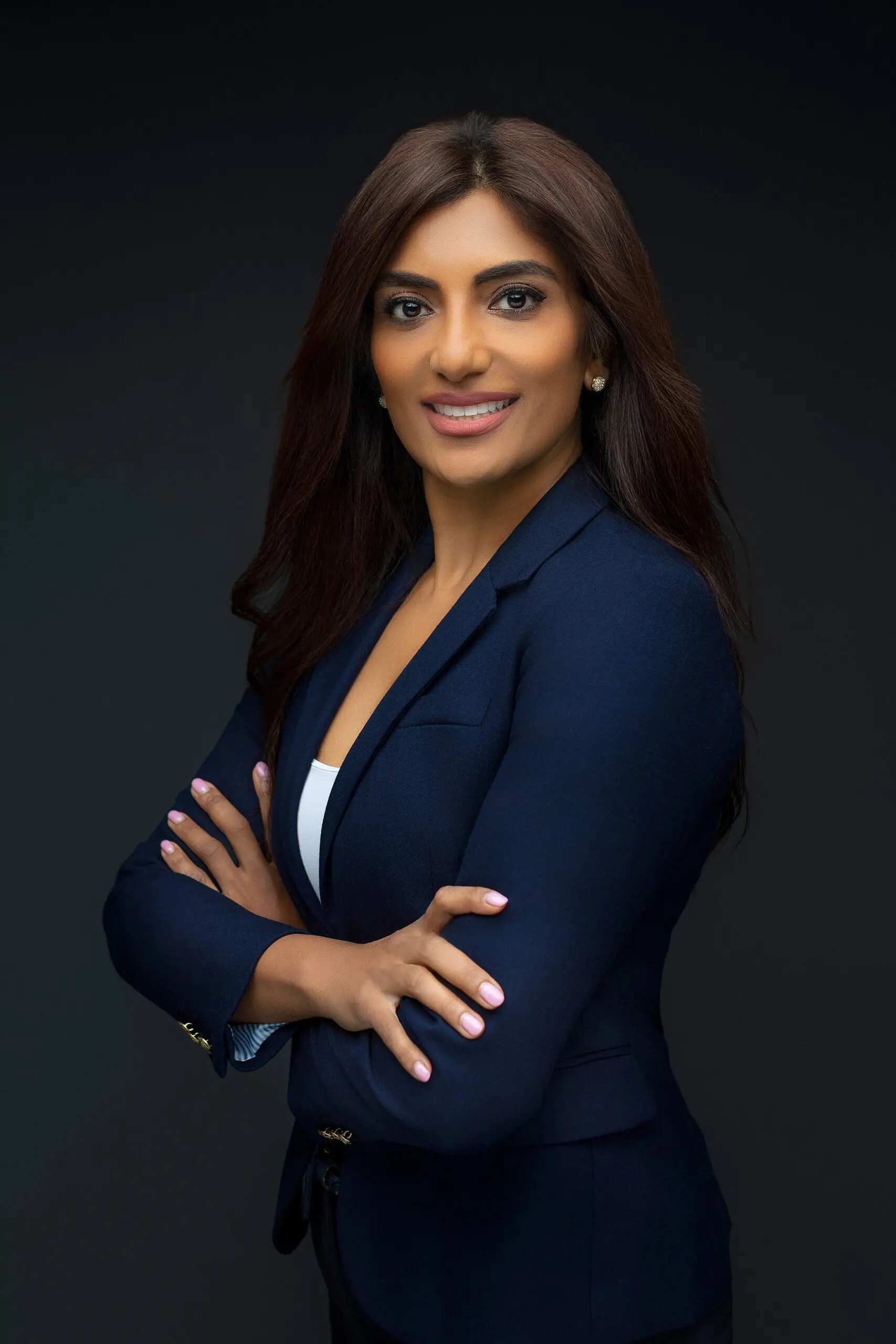 A woman with long dark hair, wearing a navy blazer and white top, stands with arms crossed and smiles confidently against a plain dark background in Connecticut.