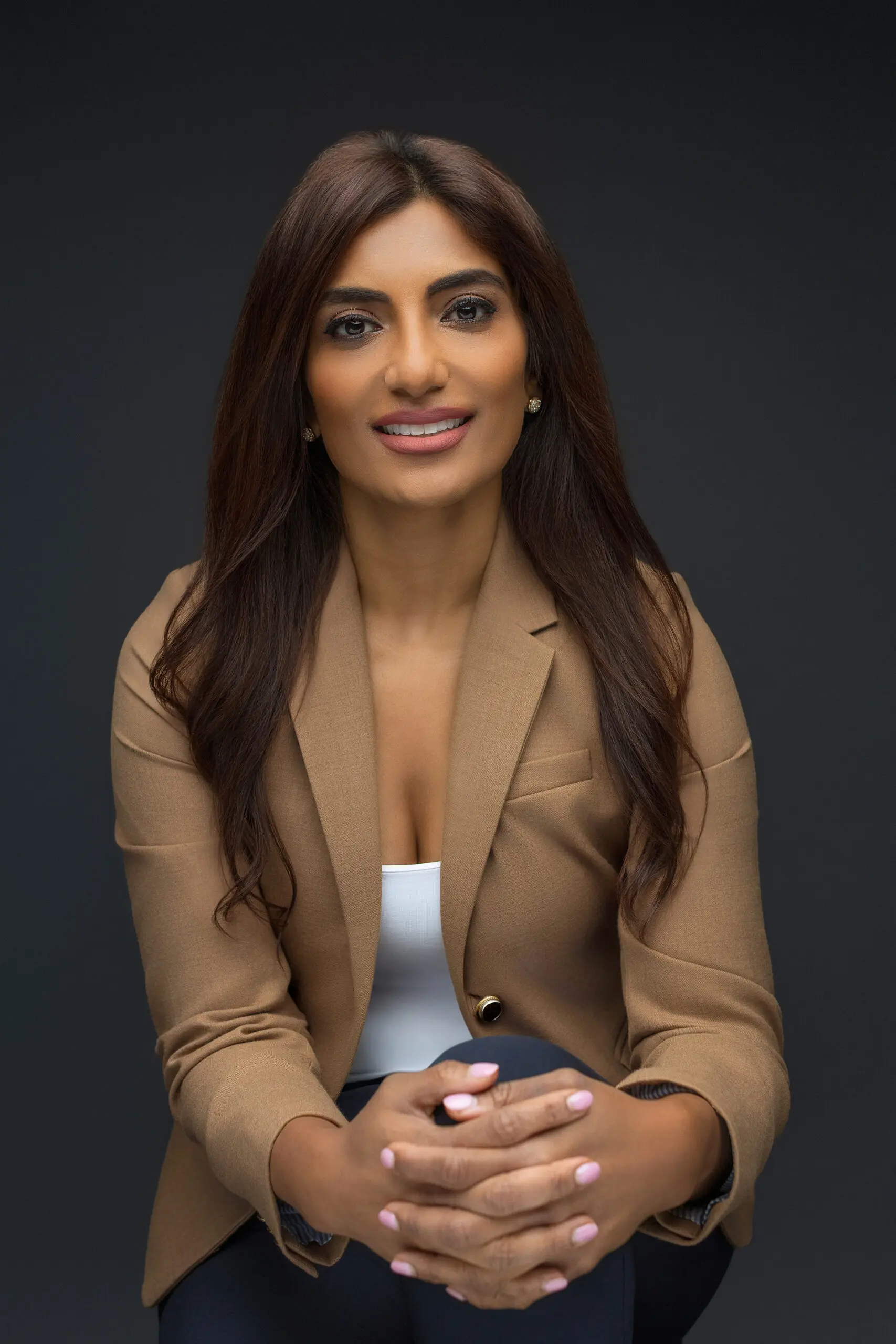 A woman with long dark hair, wearing a tan blazer and white top, sits with her hands clasped and smiles at the camera against a dark background having her professional photos taken in CT.