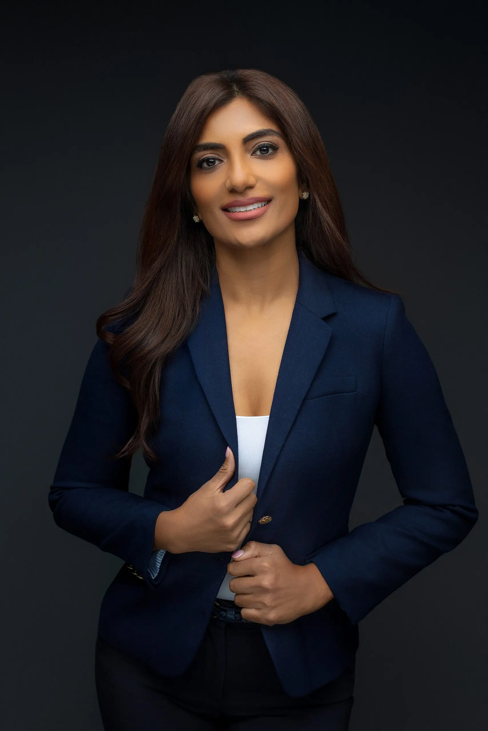 A woman with long dark hair wearing a navy blazer and white top poses confidently against a dark background, smiling slightly with one hand holding her blazer for her professional headshots in connecticut.