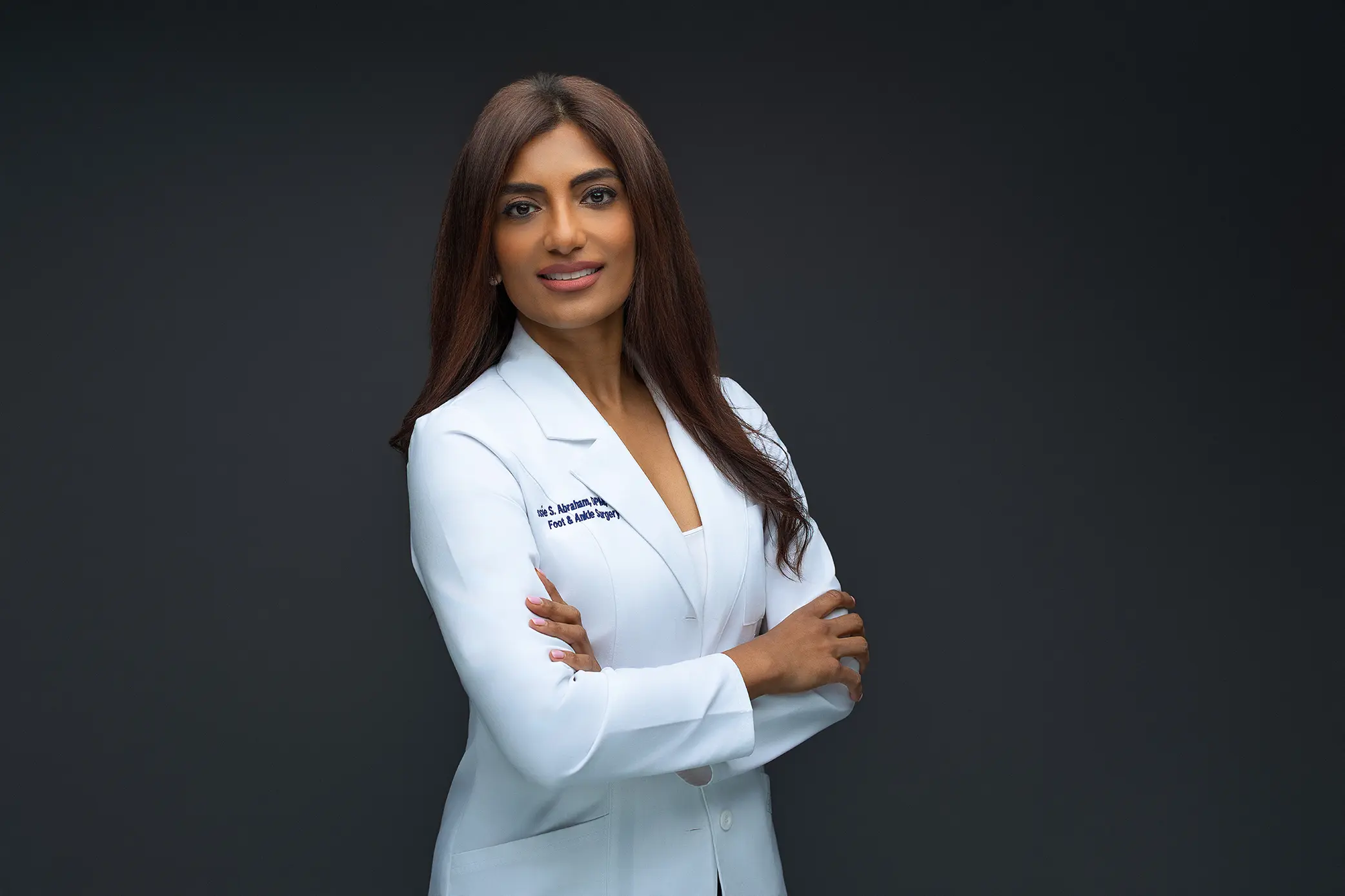 A confident woman with long brown hair wearing a white medical coat stands against a dark gray background, smiling with her arms crossed during her connecticut headshot photos.
