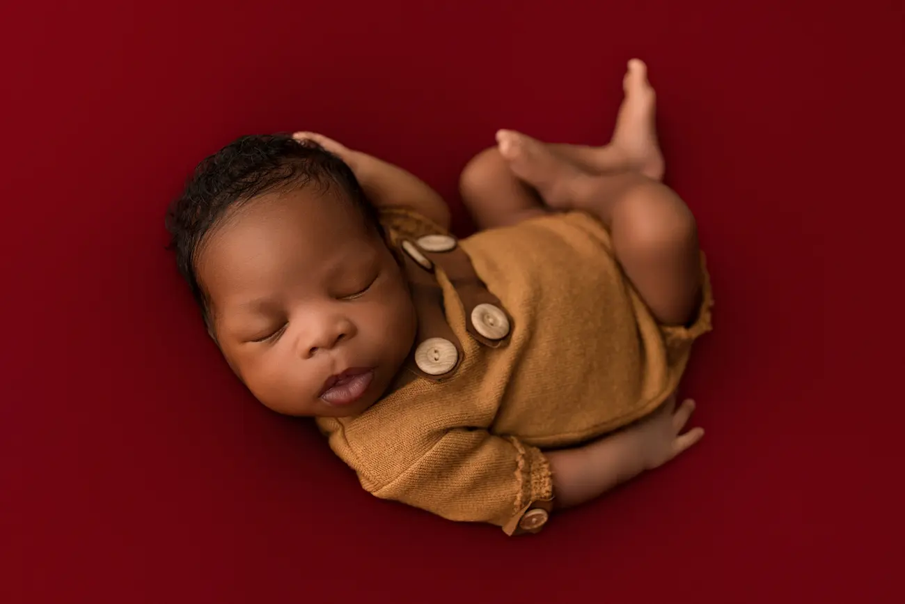 A newborn baby wearing a brown outfit with large buttons sleeps peacefully on a red background.