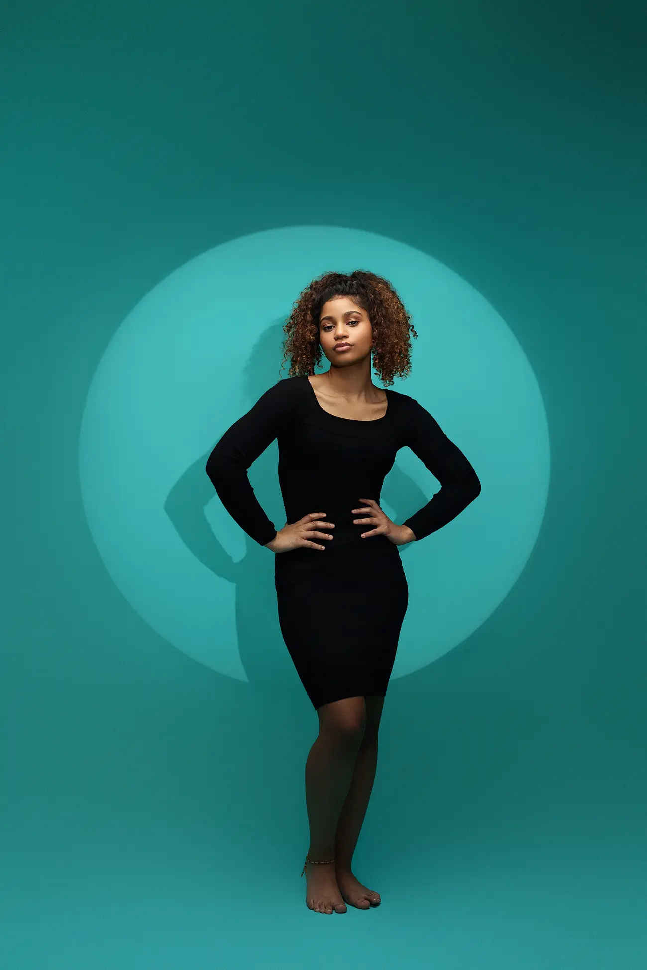 A woman with curly hair stands barefoot on a teal floor, wearing a fitted black dress. She poses confidently with hands on hips in front of a teal wall, illuminated by a circular spotlight, captured by a skilled headshot photographer Connecticut.