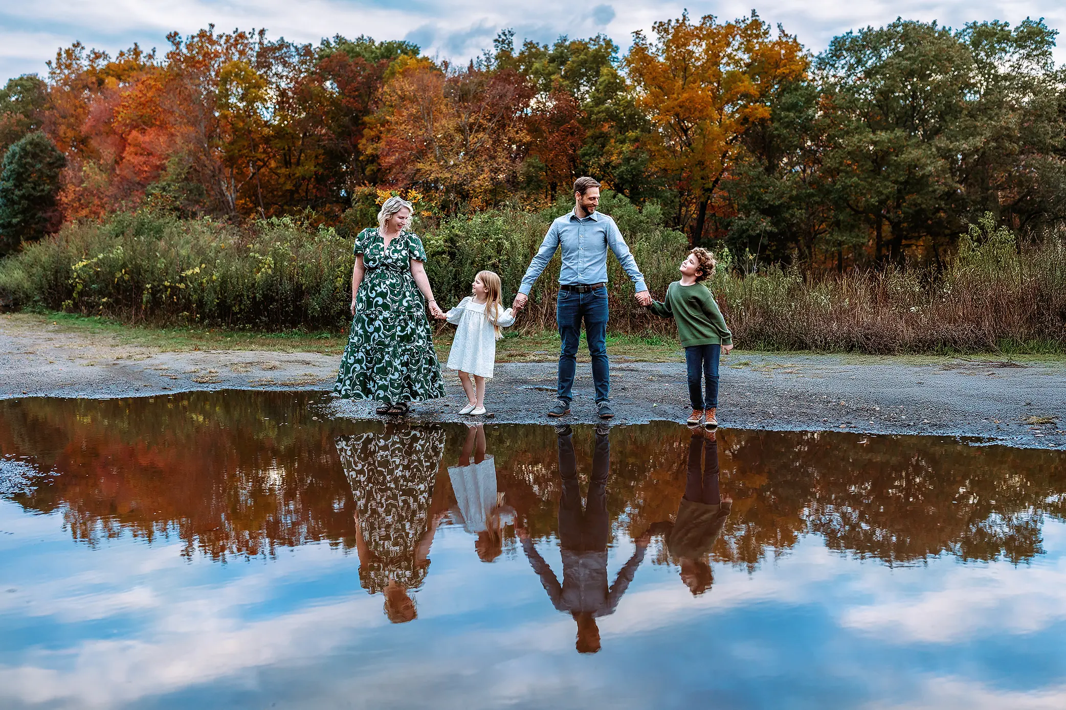 A family of four, holding hands, stands by a large puddle reflecting their image. Behind them are colorful autumn trees and a cloudy sky. The adults and two children smile together outdoors.