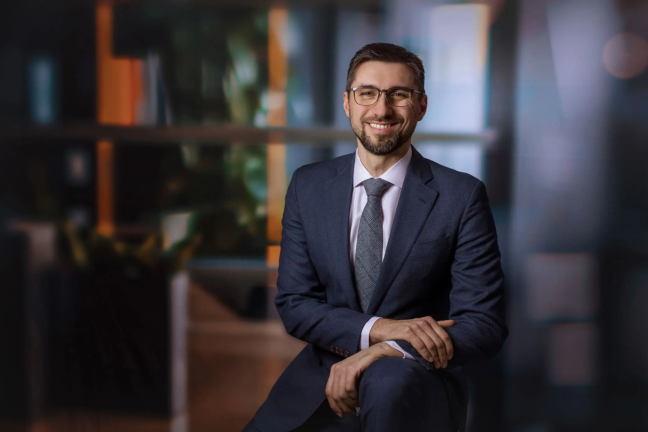 A man in a blue suit, white shirt, and patterned tie sits with a smile against a blurred office background. He has short hair, glasses, and a beard. The atmosphere is professional and warm in a connecticut studio.