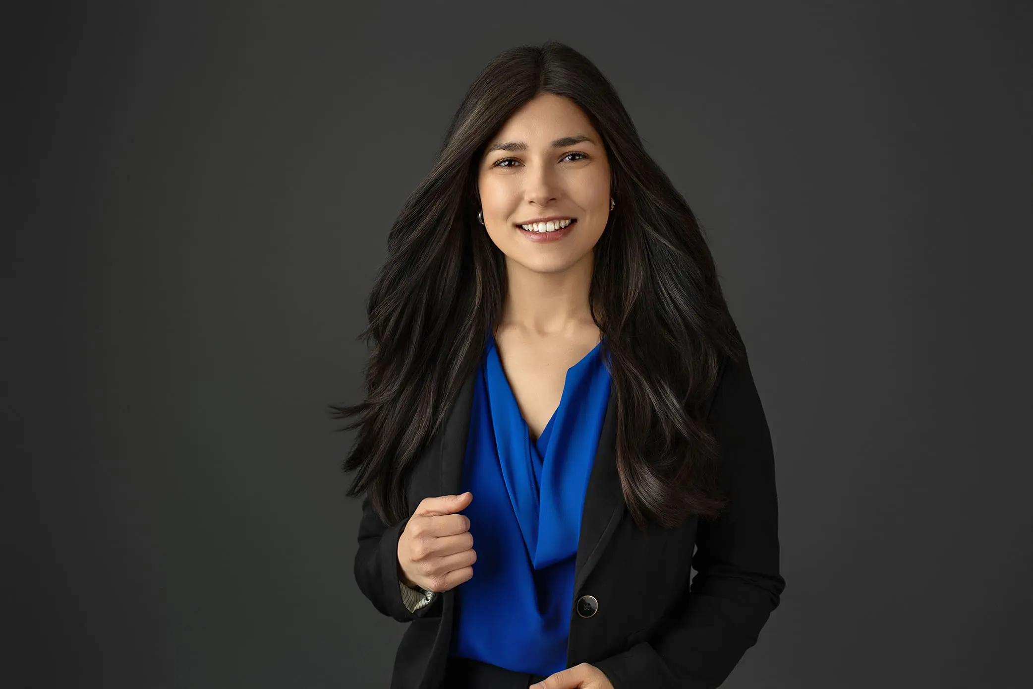 A woman with long dark hair smiles while posing against a gray background. She wears a bright blue blouse under a black blazer for her headshot photograph. Captured by Headshot Photographer in Connecticut.