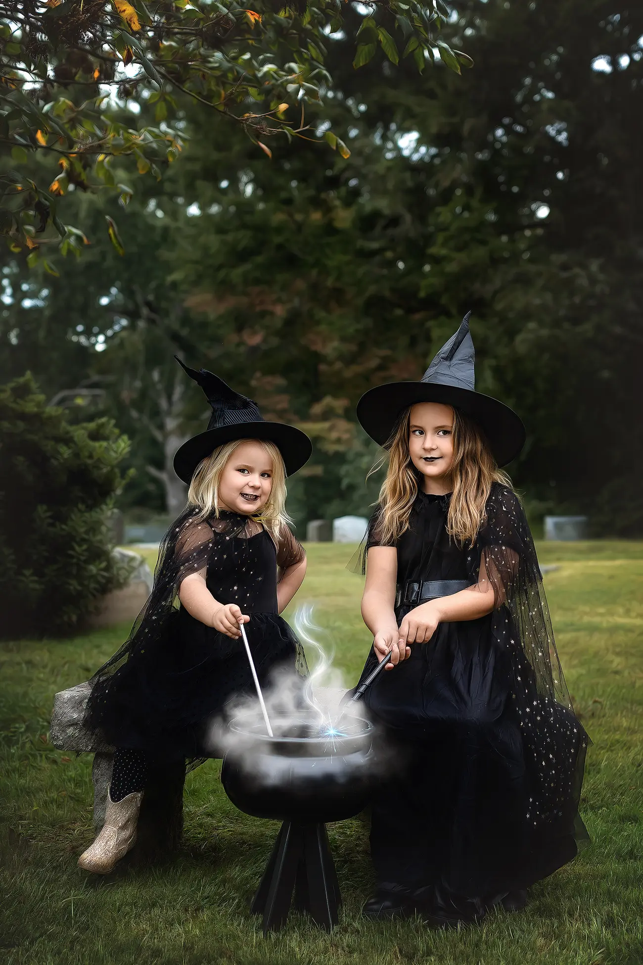 Two young girls dressed as witches sit outdoors, wearing black hats and capes. They stir a black cauldron with sticks, with white smoke rising from it, and smile at the camera. Lush greenery surrounds them.