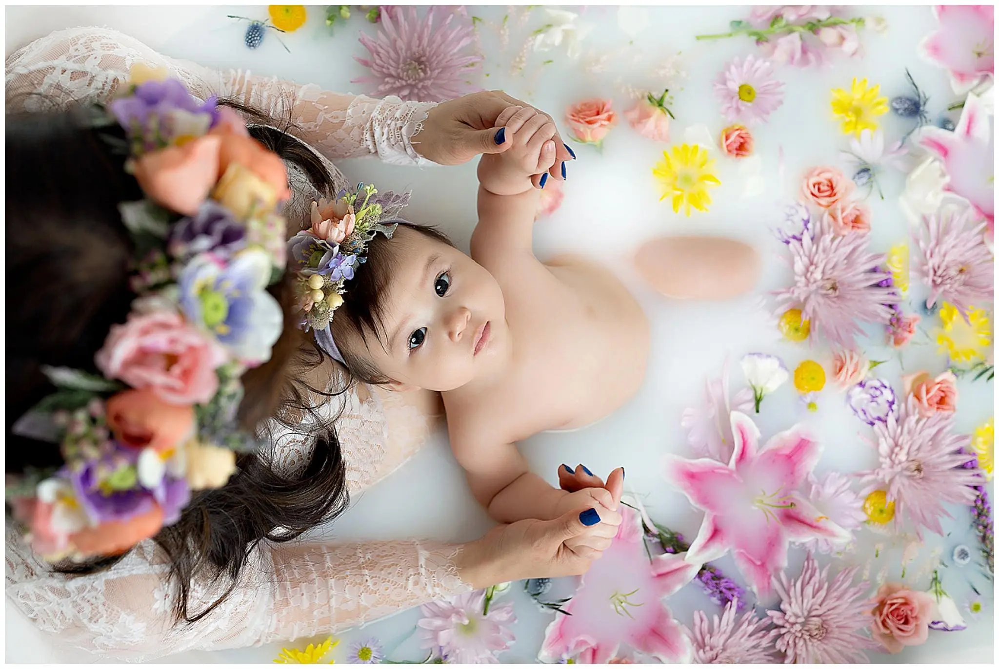 A baby lying in a floral milk bath, held by an adult with painted nails. The adult wears a floral crown. The tub is filled with colorful flowers, including lilies, daisies, and roses, floating on the surface.
