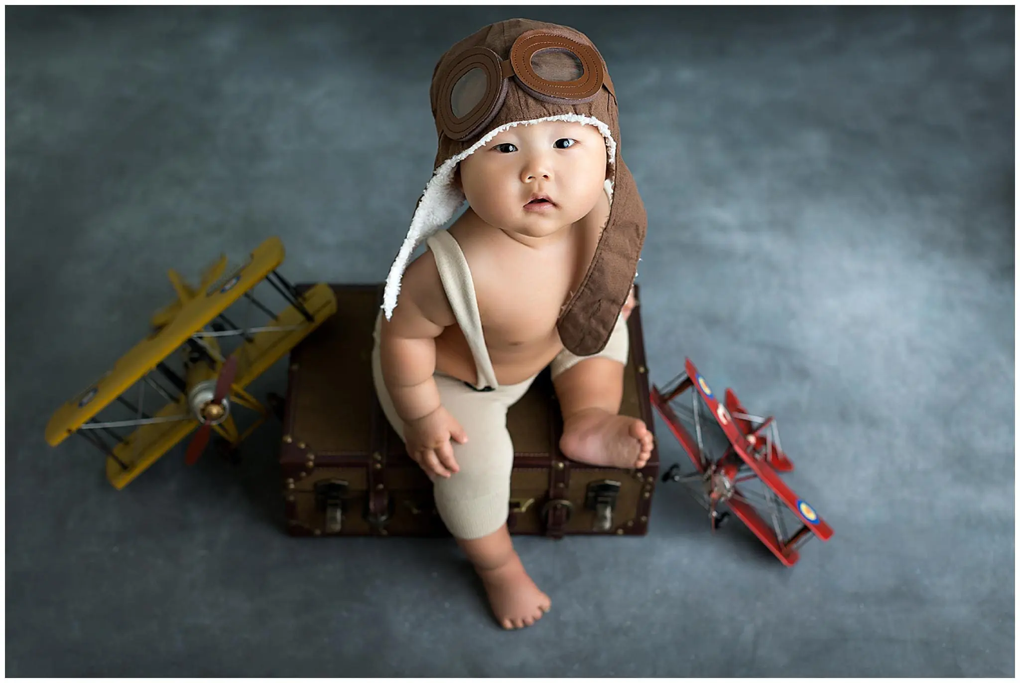 A baby photographer captures a charming scene: a baby in an aviator hat sits on a suitcase amidst toy airplanes. The little one gazes upward, with two toy biplanes, one yellow and one red, resting cheerfully on the floor nearby.