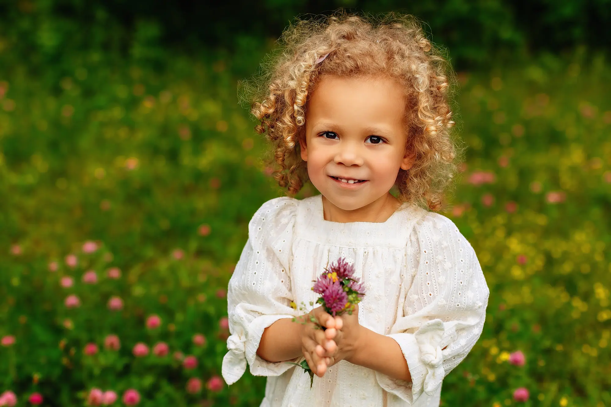 A young child with curly blond hair, wearing a white dress, stands in a field of wildflowers and smiles while holding a small bouquet of purple flowers. The background is green and vibrant. Perfect for her Connecticut family photos.