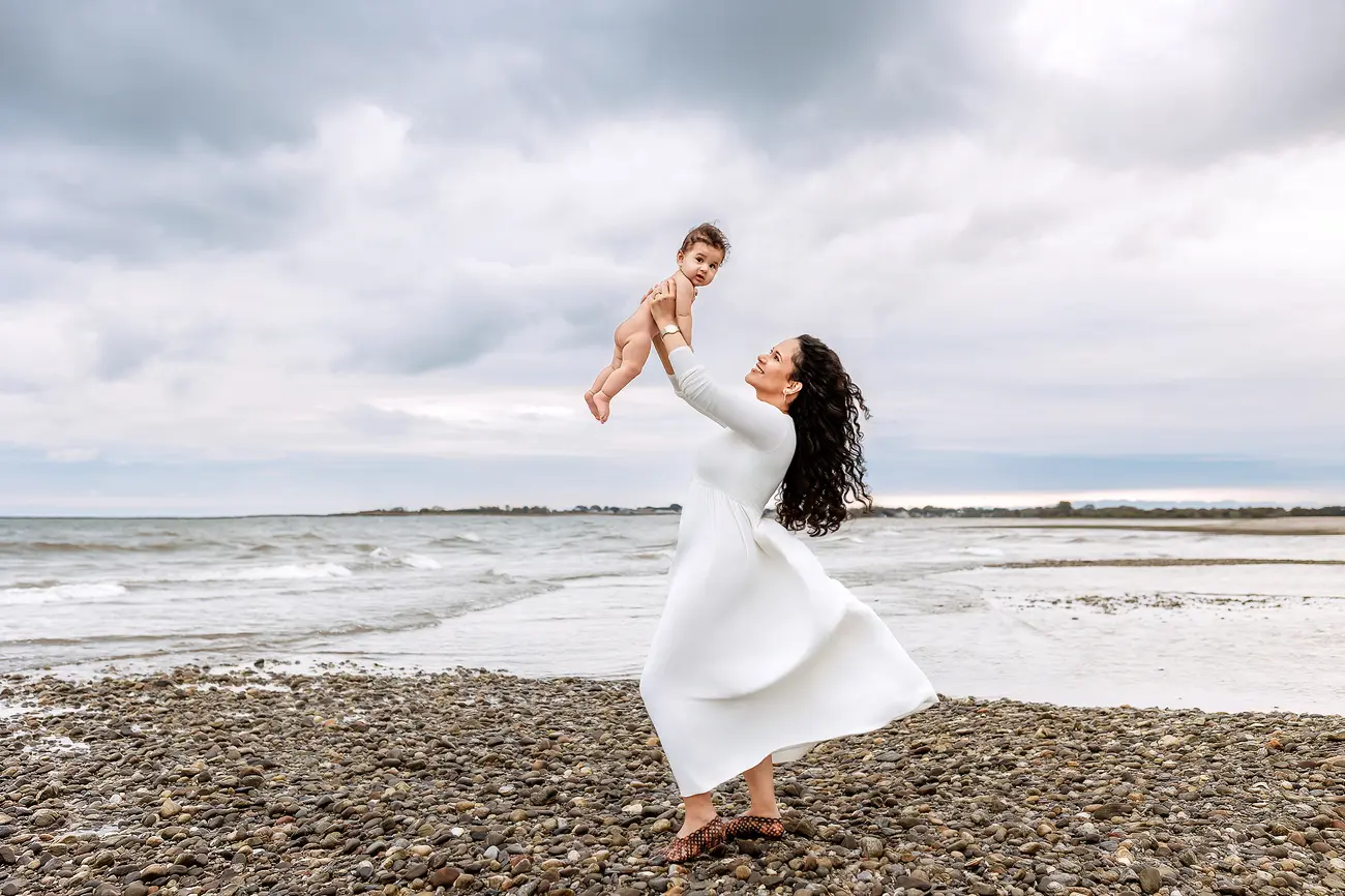 A woman in a white dress lifts a smiling baby above her head while standing on a rocky beach, with waves and a cloudy sky in the background.