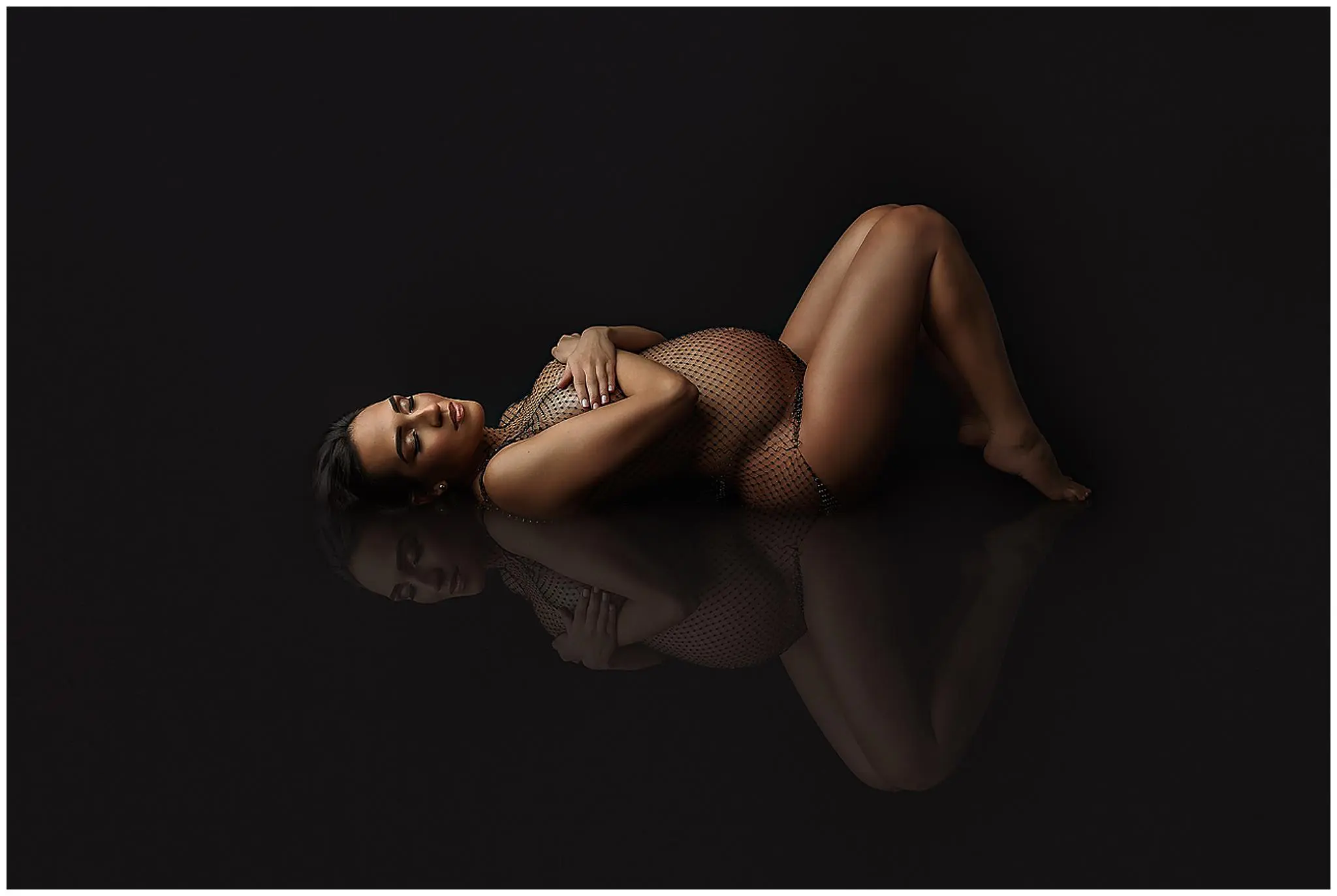A pregnant woman in a mesh bodysuit reclines on her side against a dark background, her arms folded gently over her chest, with her reflection visible beneath her in Milford CT.