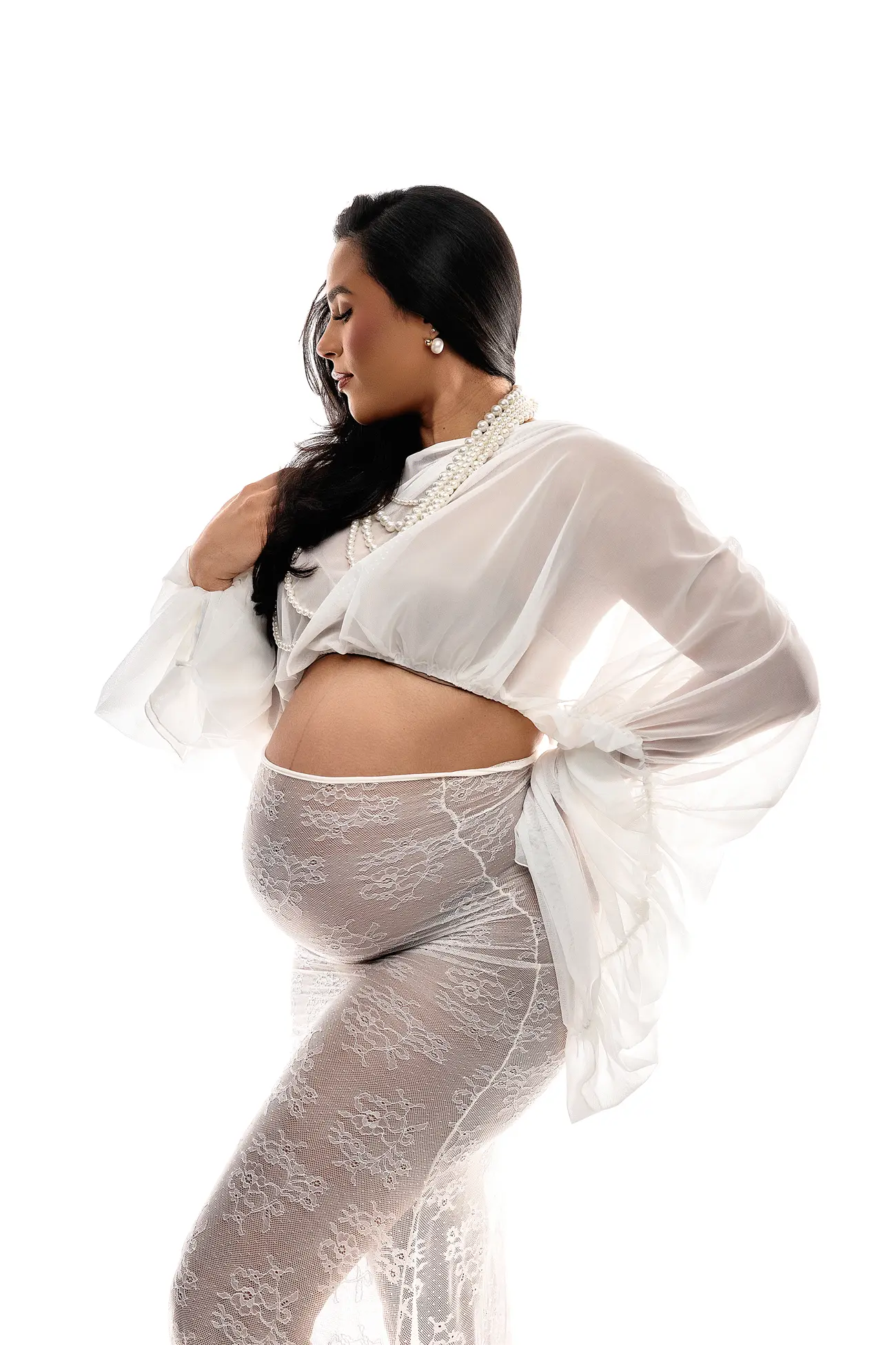 A pregnant woman in a white lace bodysuit and sheer top poses against a white background.