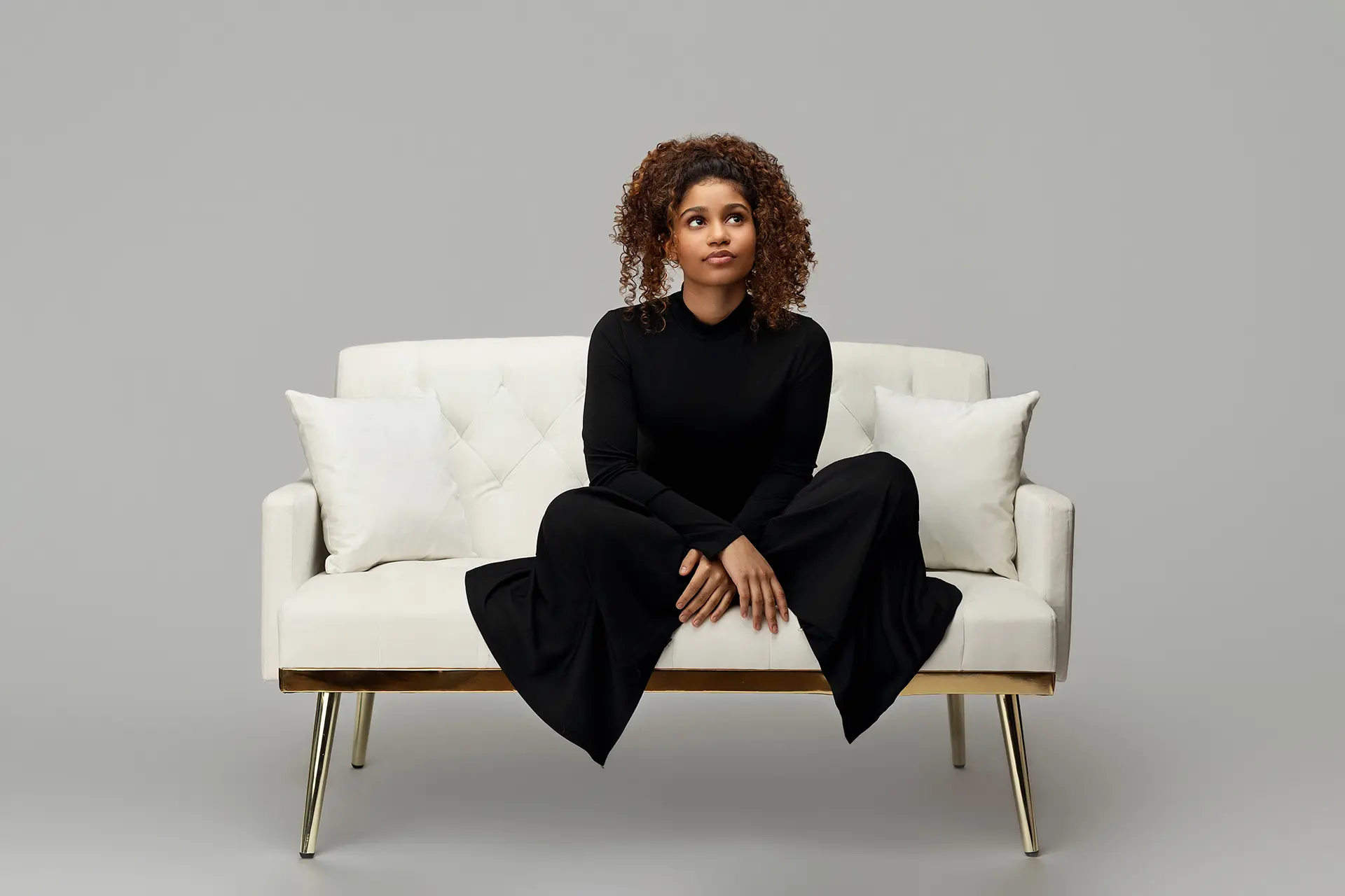 A woman with curly hair, wearing a long black dress, sits on a modern white sofa with two white pillows against a plain gray background. She looks slightly upward with a thoughtful expression.