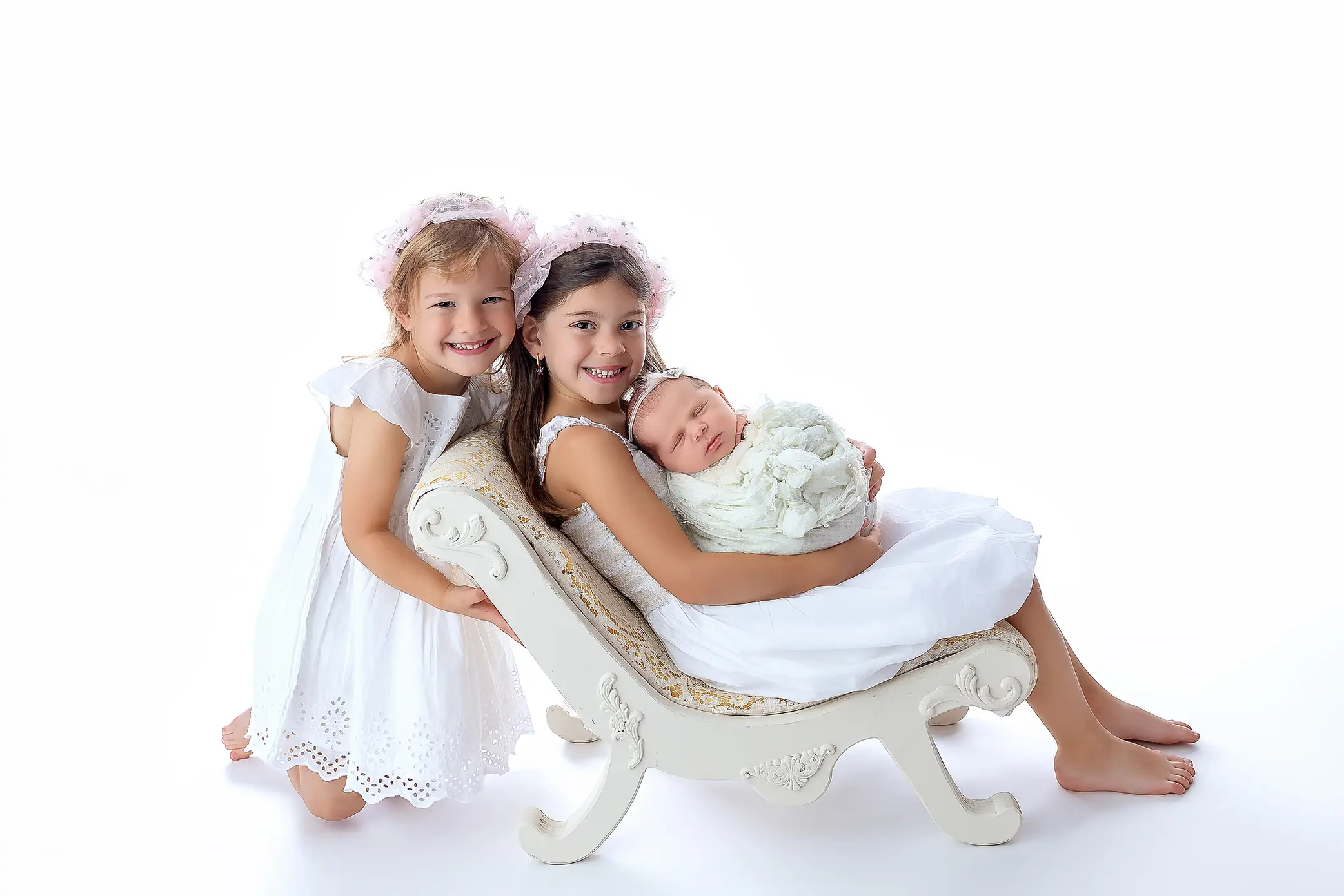 Two young girls in white dresses and bonnets pose with a newborn wrapped in a blanket on a small ornate chaise lounge, all smiling against a white background.